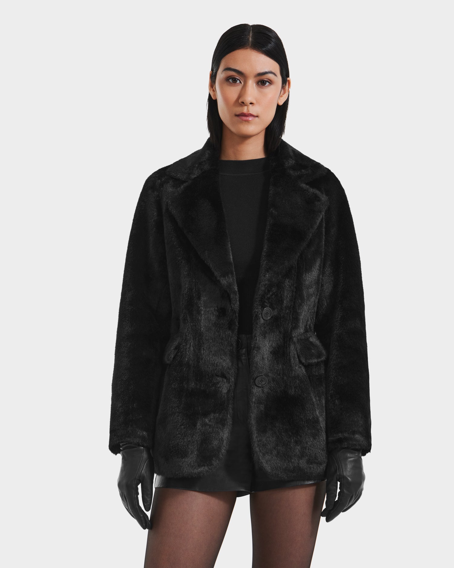 A woman with straight dark hair models the Rudsak ASPEN F - 8125604 Black faux fur jacket, paired with black gloves, shorts, and top against a white background—an effortlessly chic cold-weather look.