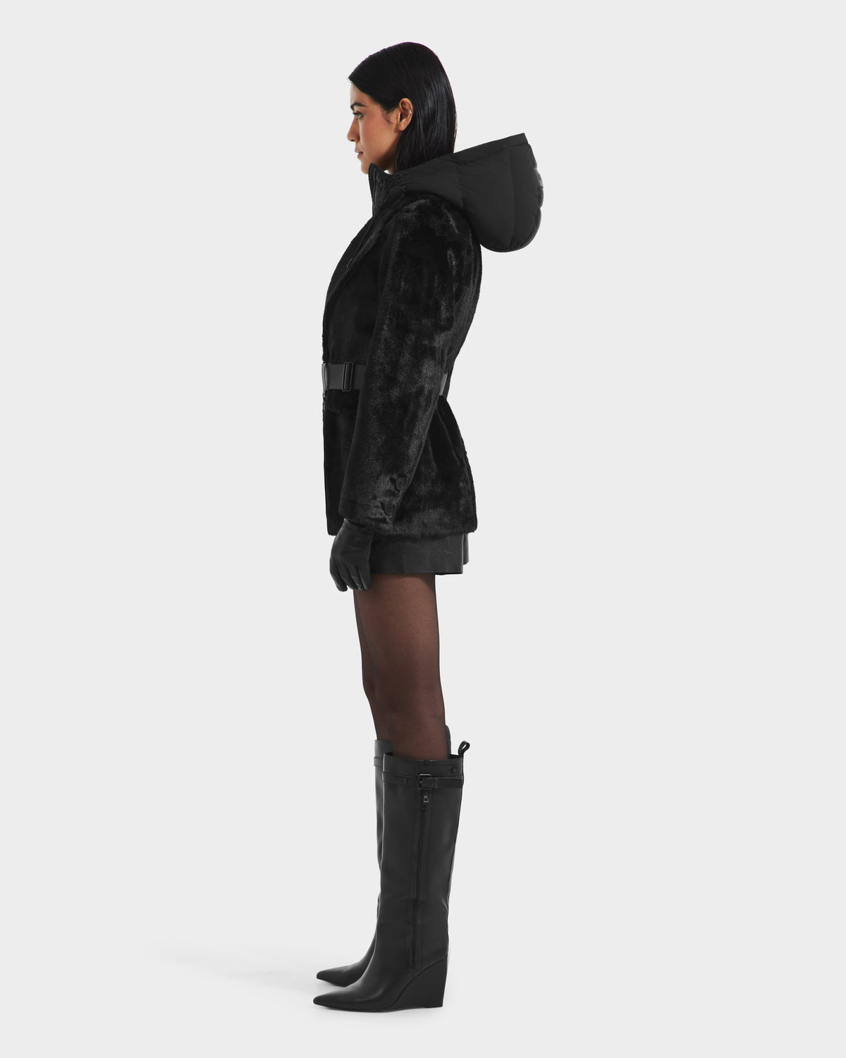 A woman in profile wears the Rudsak ASPEN F - 8125604 Black faux fur jacket with a down-insulated hood, paired with black gloves, shorts, tights, and knee-high boots—an essential cold-weather look against a white background.
