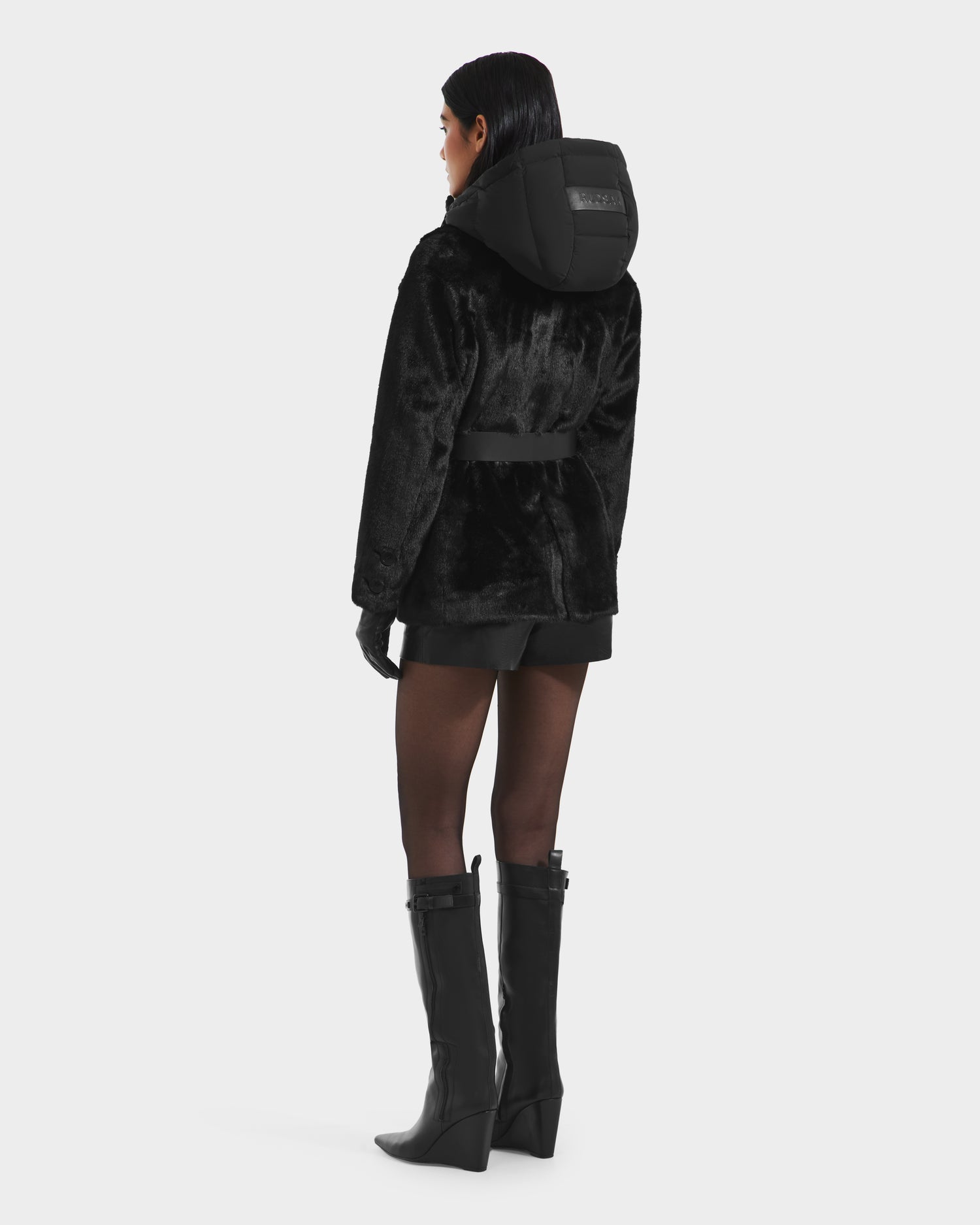 A woman faces away, wearing the Rudsak ASPEN F - 8125604 Black faux fur jacket with a down-insulated hood, styled with black shorts, gloves, and knee-high boots—her sleek hair pops against the white backdrop for a chic winter look.