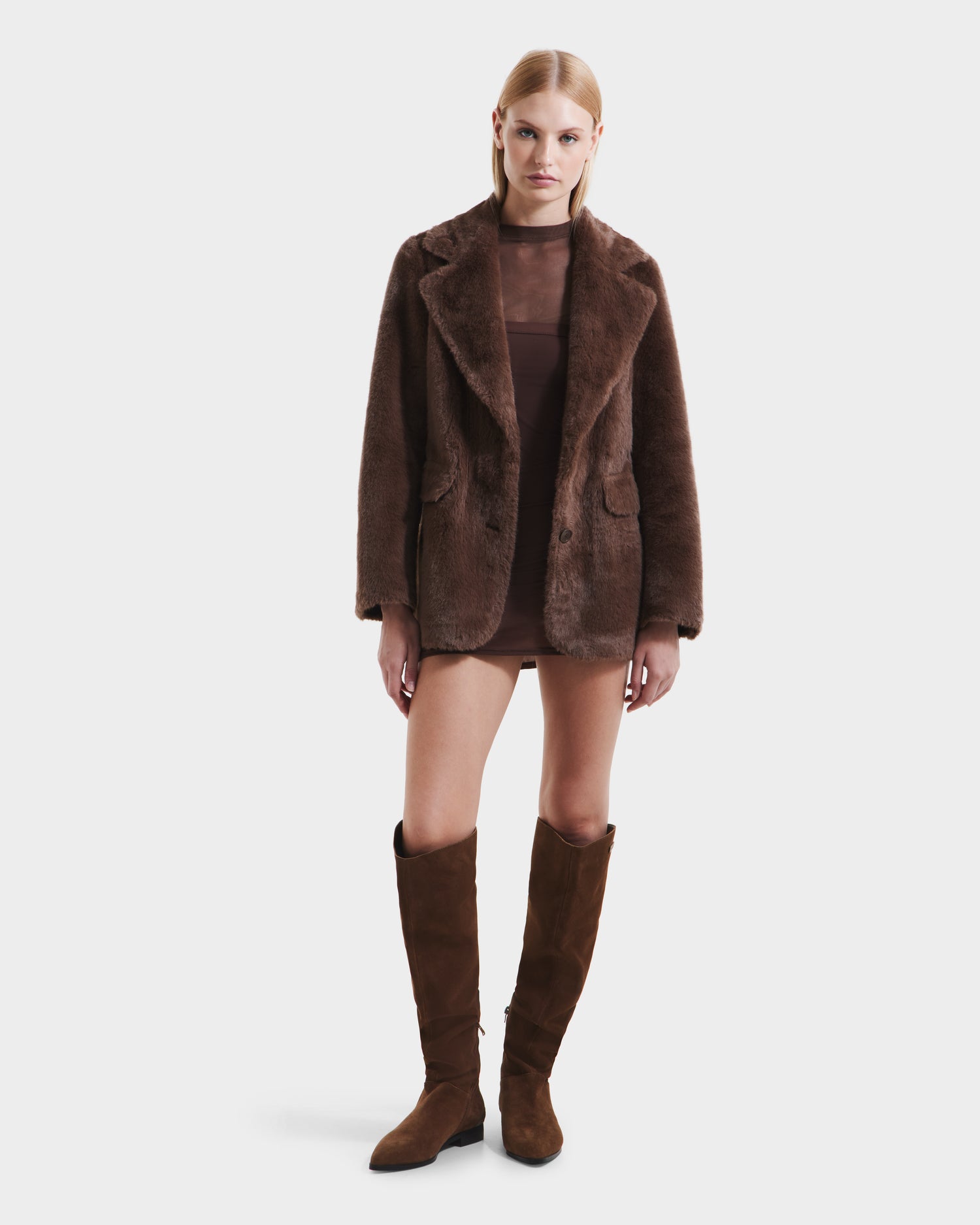 A woman faces forward wearing the Rudsak ASPEN F - 8125604 Mocha faux fur jacket, styled over a sheer brown mini dress and knee-high suede boots, as part of her cold-weather look against a plain white background.