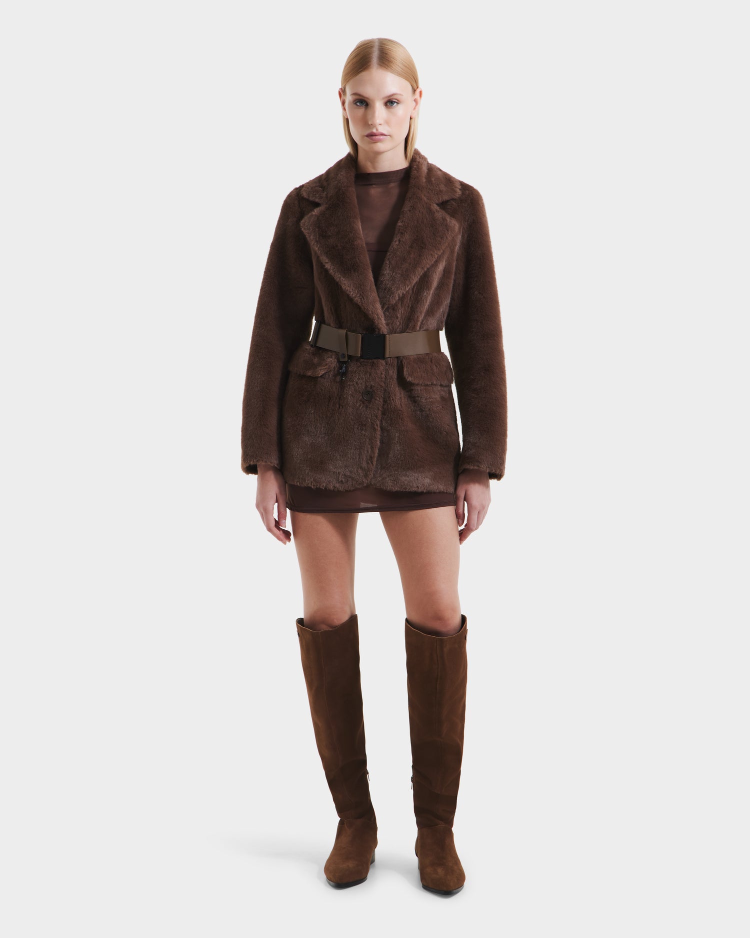 A woman faces forward in the Rudsak ASPEN F - 8125604 Mocha faux fur jacket with belt, brown mini dress, and knee-high brown suede boots—a chic cold-weather look against a plain white background.