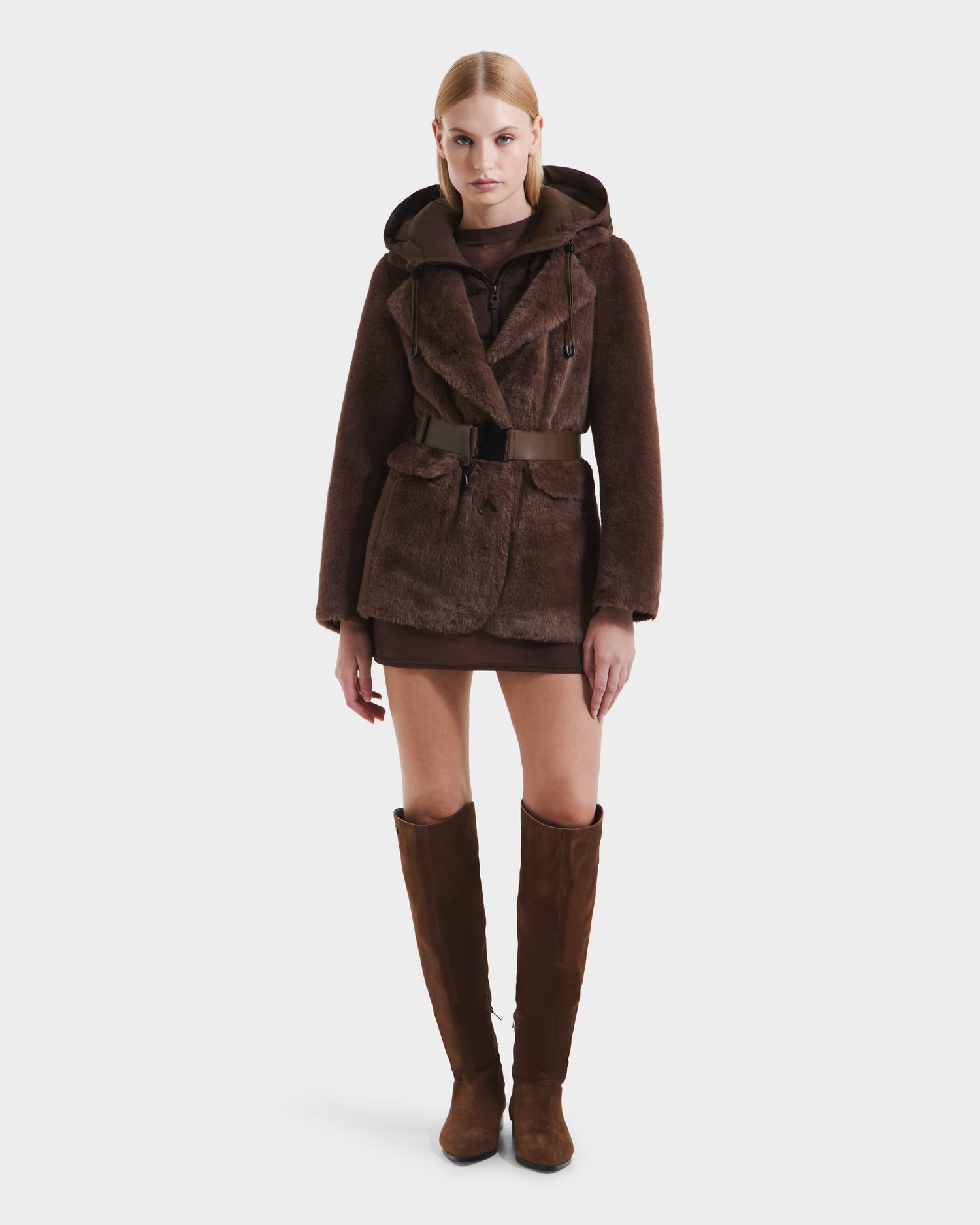 A woman with straight blonde hair and a neutral expression wears the Rudsak ASPEN F - 8125604 Mocha faux fur jacket with removable down-insulated hood, belted waist, over a brown dress and tall brown suede boots against a white background.