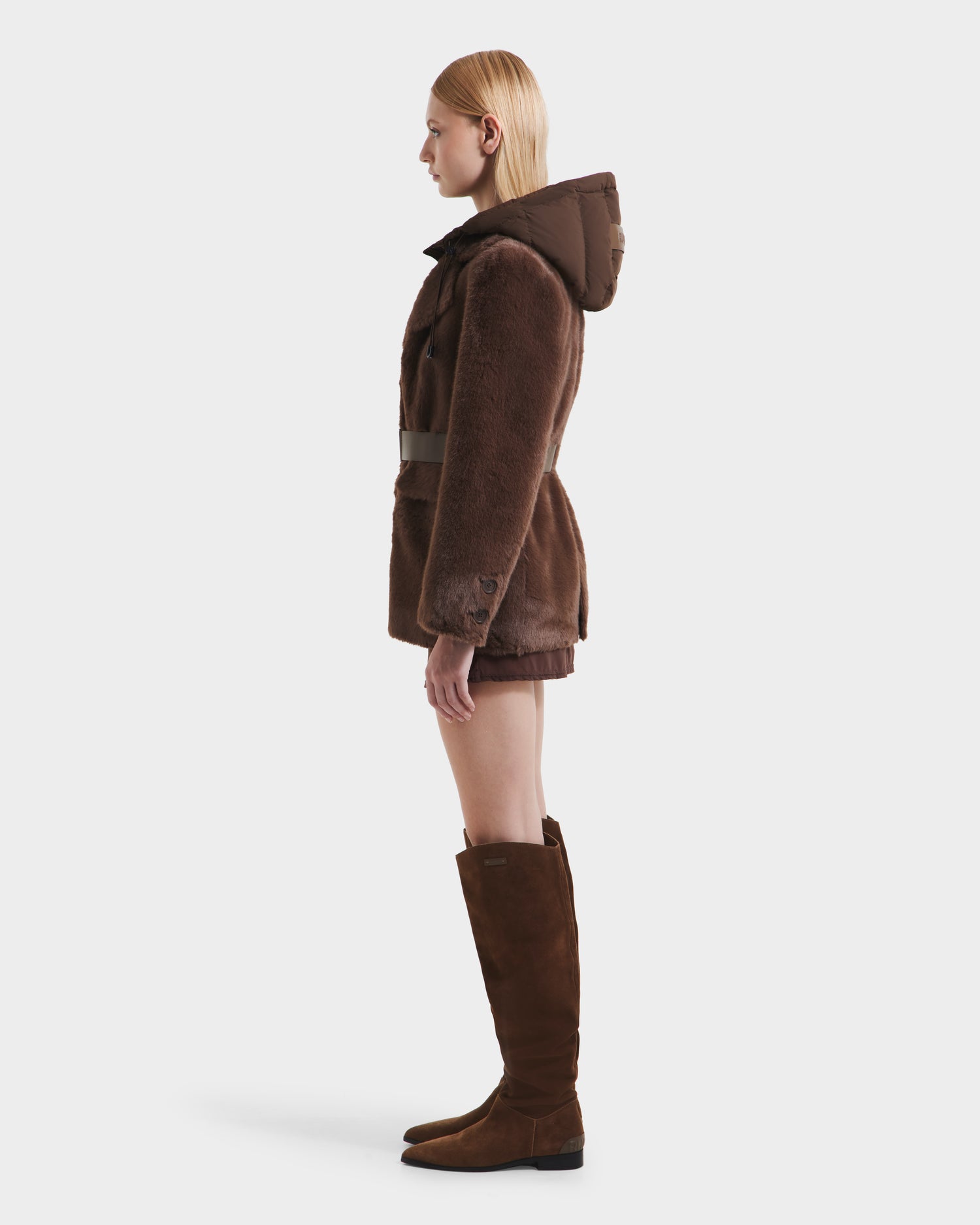 A woman with straight blonde hair stands in profile, wearing the Rudsak ASPEN F - 8125604 Mocha women’s faux fur jacket with a belted waist—an effortlessly chic cold-weather look. She pairs it with knee-high brown suede boots against a plain white background.