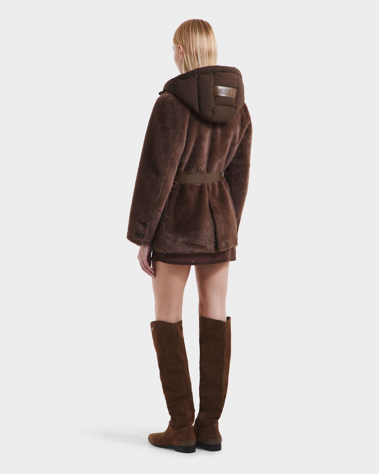 A person with straight blonde hair is seen from the back wearing the Rudsak ASPEN F - 8125604 Mocha women’s faux fur jacket with a belted waist and removable down-insulated hood, paired with brown knee-high boots, against a plain white background.