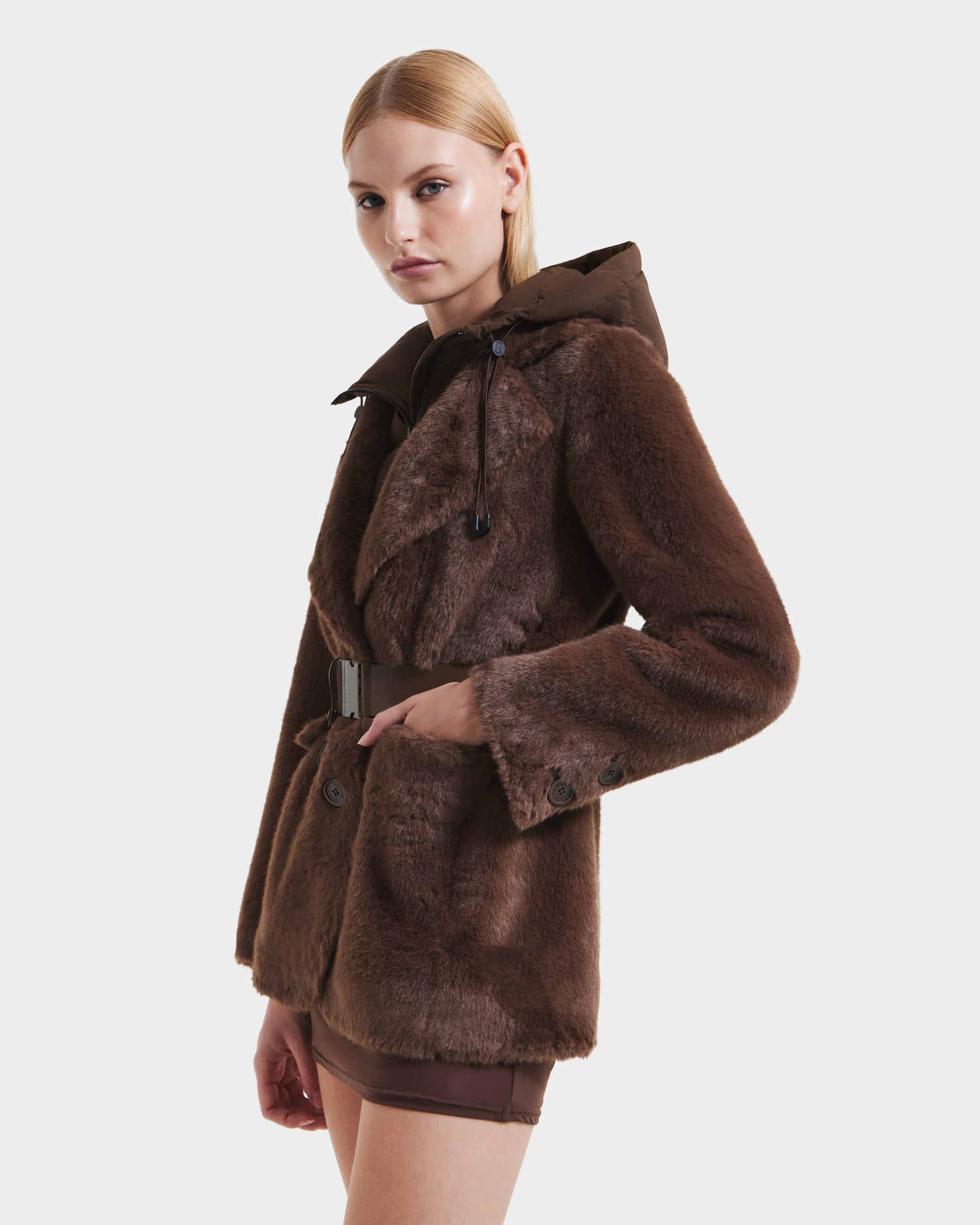 A woman with straight blonde hair models the Rudsak ASPEN F - 8125604 Mocha women’s faux fur jacket, featuring large front pockets, a belt, and a removable down-insulated hood. She pairs it with brown shorts against a plain white background.