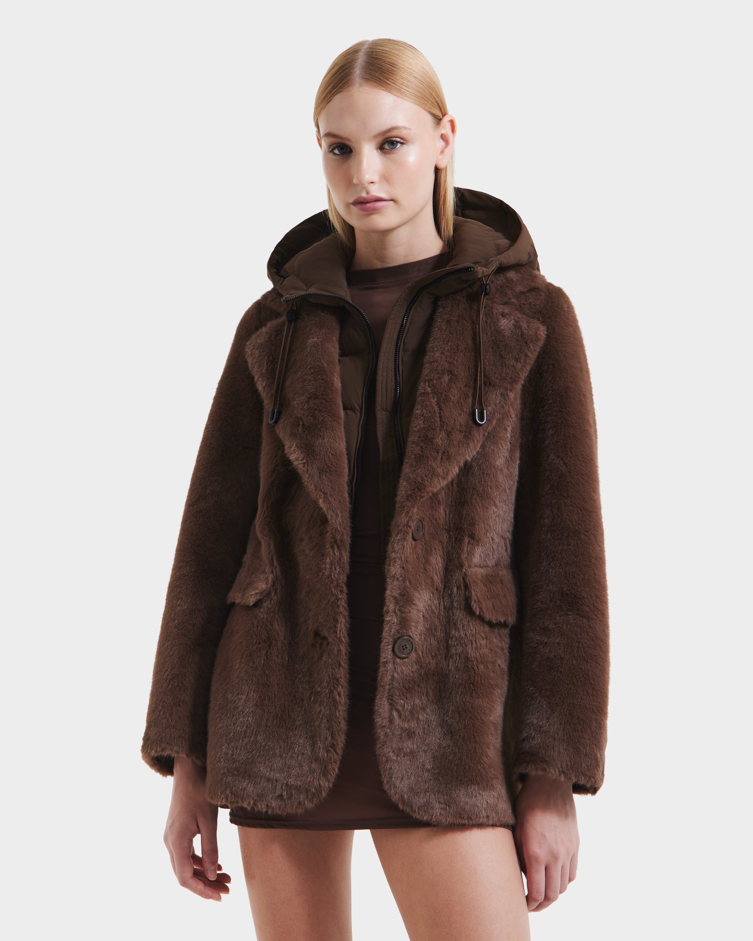 A woman with straight blonde hair models the Rudsak ASPEN F - 8125604 Mocha women’s faux fur jacket with a removable down-insulated hood over a short brown dress, standing against a plain white background—perfect for cold weather.