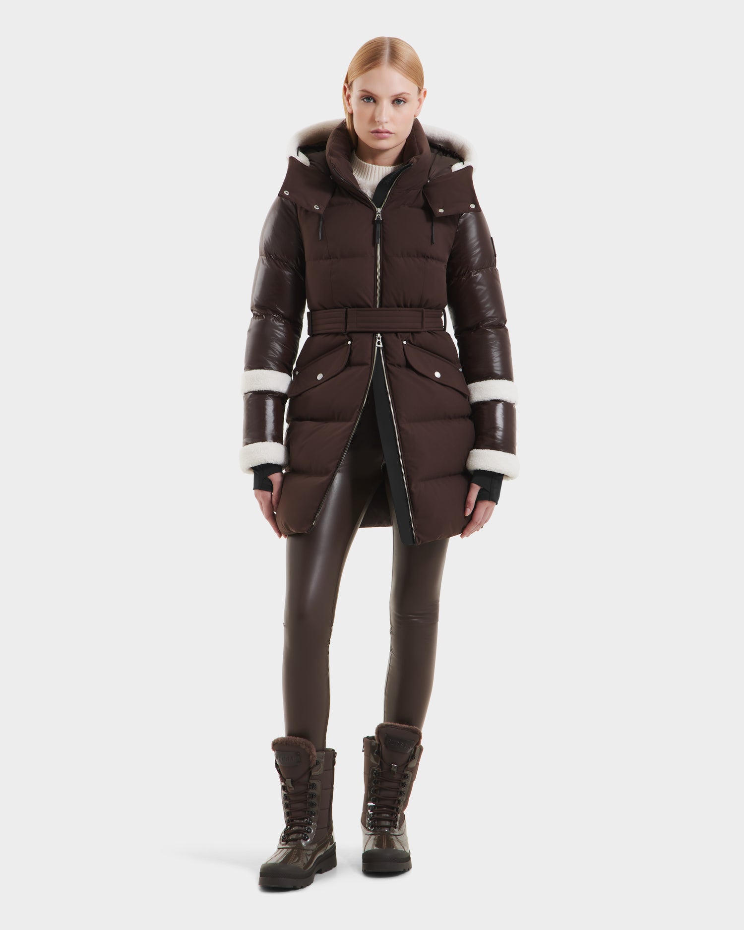 A woman faces forward in the Rudsak AUDREY L - 8124585 Espresso, a belted knee-length RDS certified down puffer with hood and white fleece trim, styled with matching brown leggings and lace-up winter boots against a plain white background.
