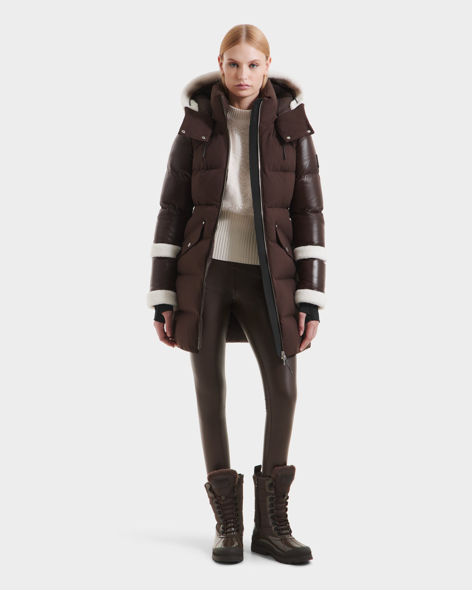 A woman faces forward against a white background wearing the Rudsak AUDREY L - 8124585 Espresso knee-length down puffer jacket with white trim, brown leather-look pants, a cream turtleneck, and brown lace-up boots.