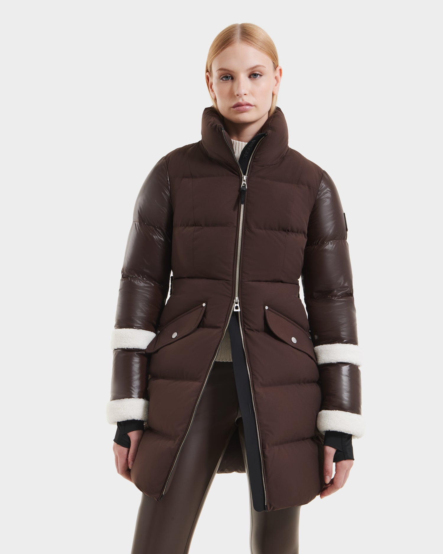 A woman wears the Rudsak AUDREY L - 8124585 Espresso, a knee-length down puffer jacket with shiny quilted sleeves and white shearling trim, made from recycled Stormshell fabric and filled with RDS-certified down. She stands against a plain white background.