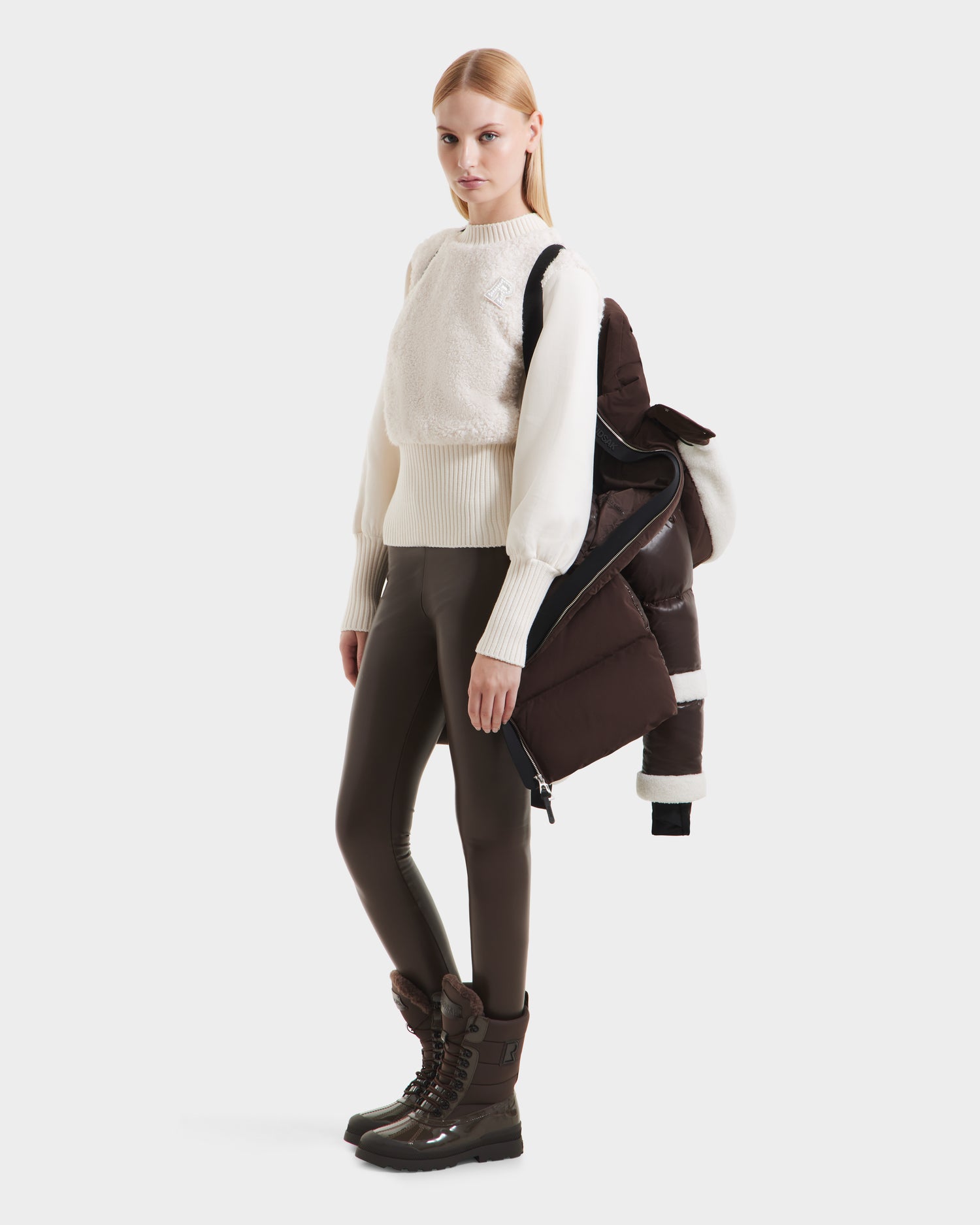 A woman faces forward in a white sweater, brown leggings, and boots, carrying the Rudsak AUDREY L - 8124585 Espresso knee-length down puffer jacket over one shoulder against a plain white background.