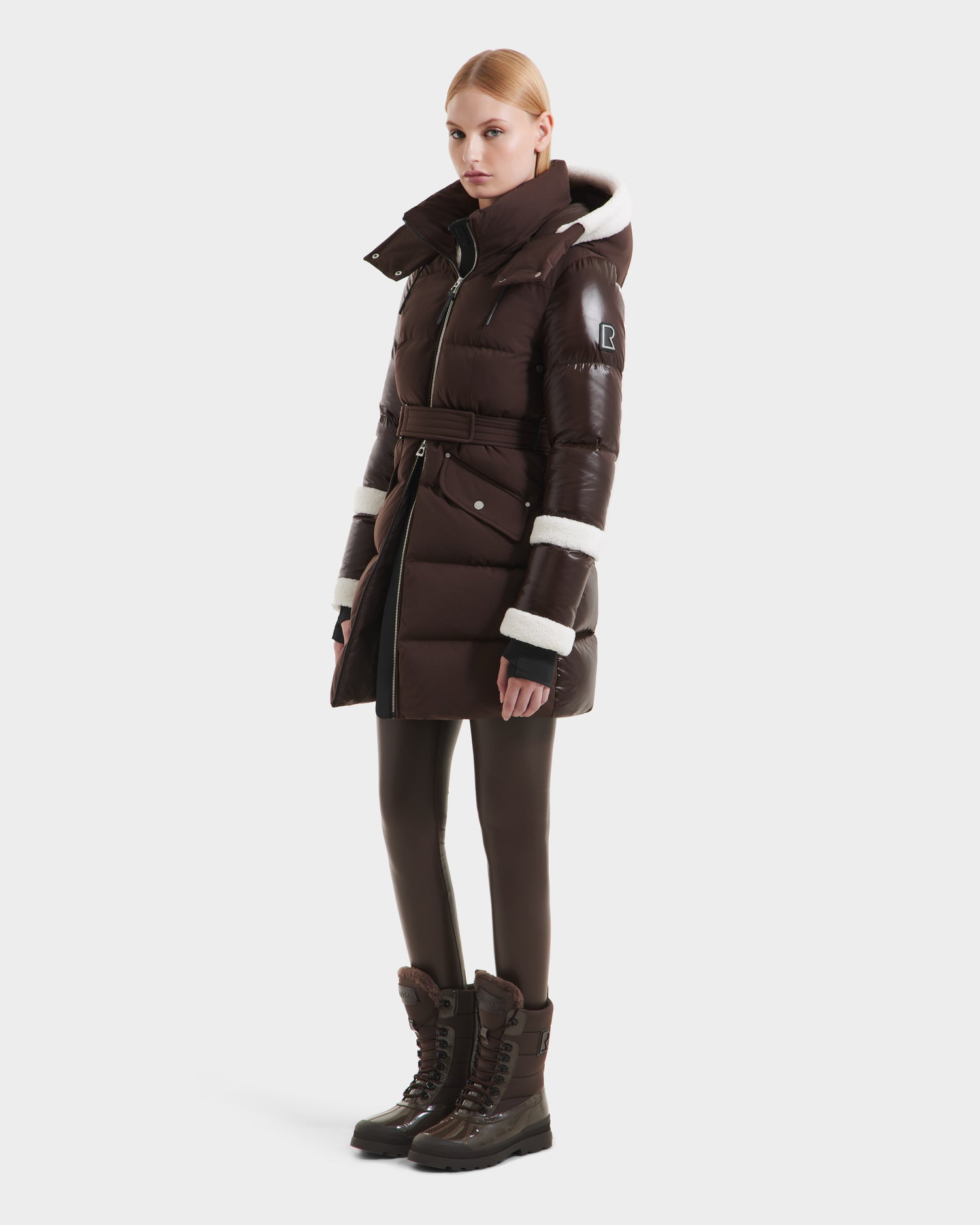 A woman models the Rudsak AUDREY L - 8124585 Espresso puffer jacket with a belted waist and high collar, paired with black leggings and brown winter boots, standing against a plain, light background.