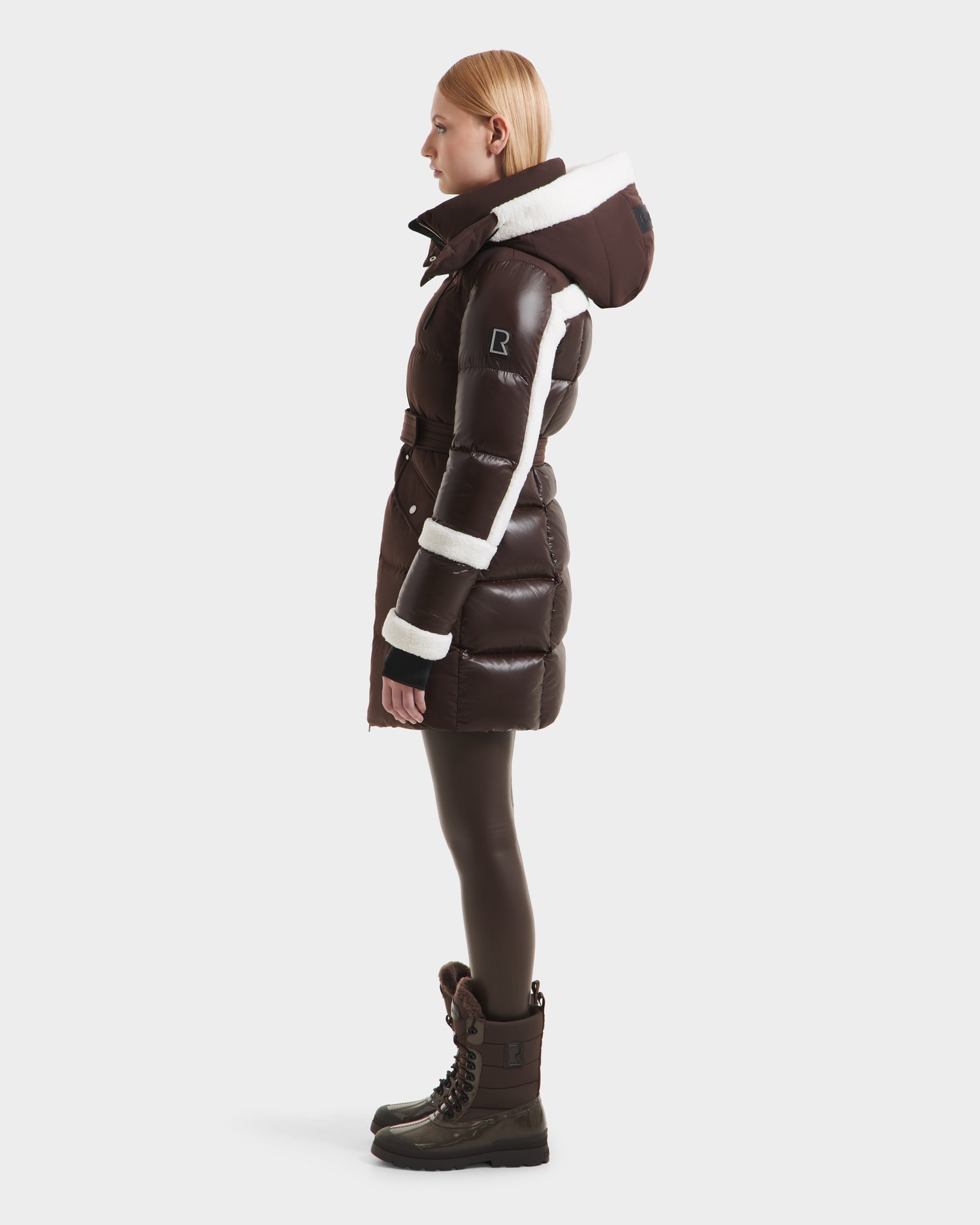 A woman with straight blonde hair wears the Rudsak AUDREY L - 8124585 Espresso, a knee-length down puffer jacket with white trim, belted waist, and hood. She pairs it with dark leggings and brown winter boots.