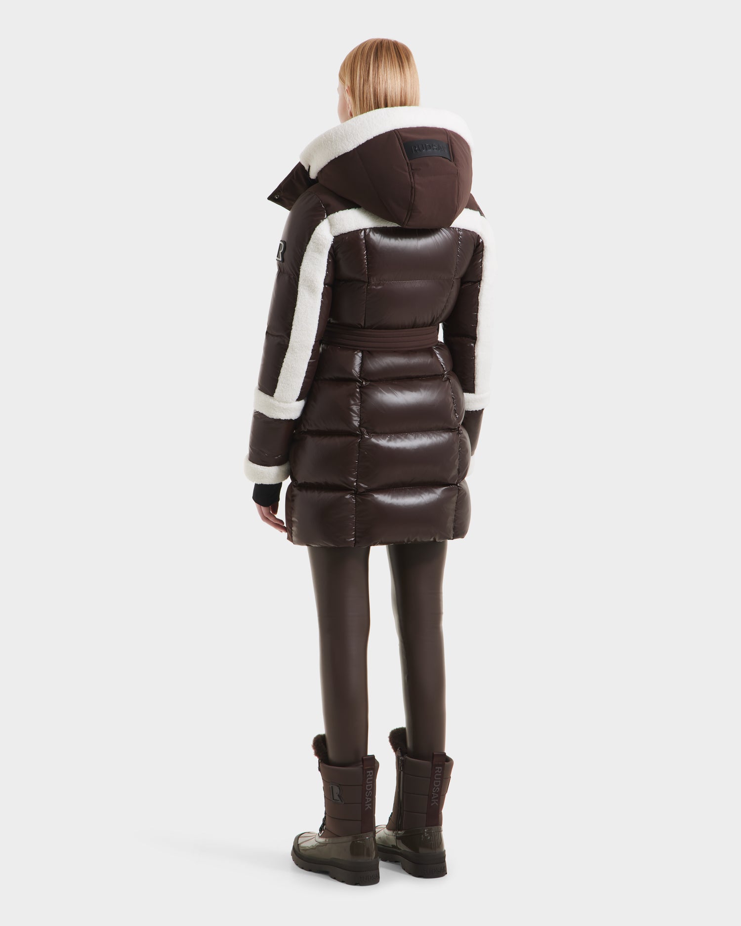 A person with straight, shoulder-length blonde hair is seen from the back wearing the Rudsak AUDREY L - 8124585 Espresso knee-length down puffer jacket with white shearling trim, paired with brown leggings and boots against a white background.