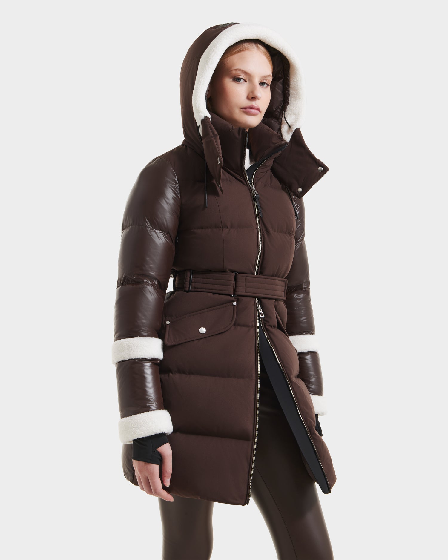 A woman wears Rudsak’s AUDREY L - 8124585 Espresso: a dark brown, knee-length puffer with RDS-certified down, shiny sleeves, a belted waist, striped details, white fleece-lined hood, and fleece-trimmed cuffs against a plain background.
