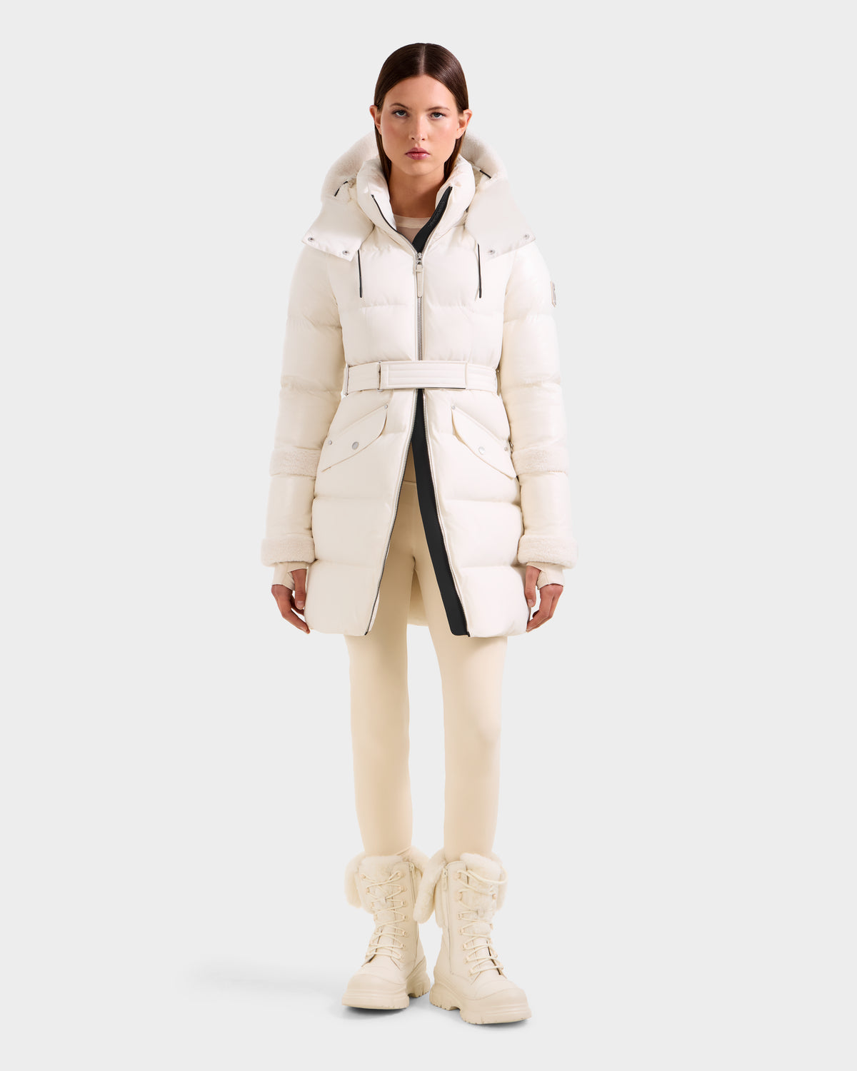 A woman wears the Rudsak AUDREY L - 8124585 Vanilla, a belted knee-length down puffer jacket with hood in recycled Stormshell fabric, paired with cream pants and matching lace-up boots, posing against a plain light gray background.
