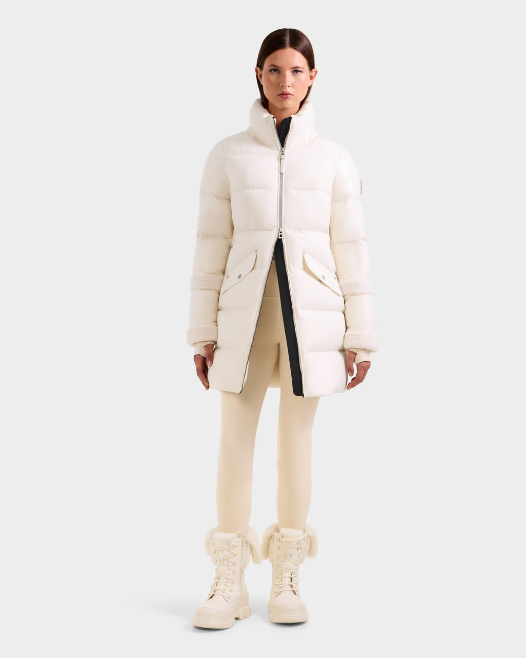 A woman faces forward wearing the Rudsak AUDREY L - 8124585 Vanilla knee-length down puffer jacket, cream leggings, and matching fur-trimmed lace-up boots against a plain light gray background.