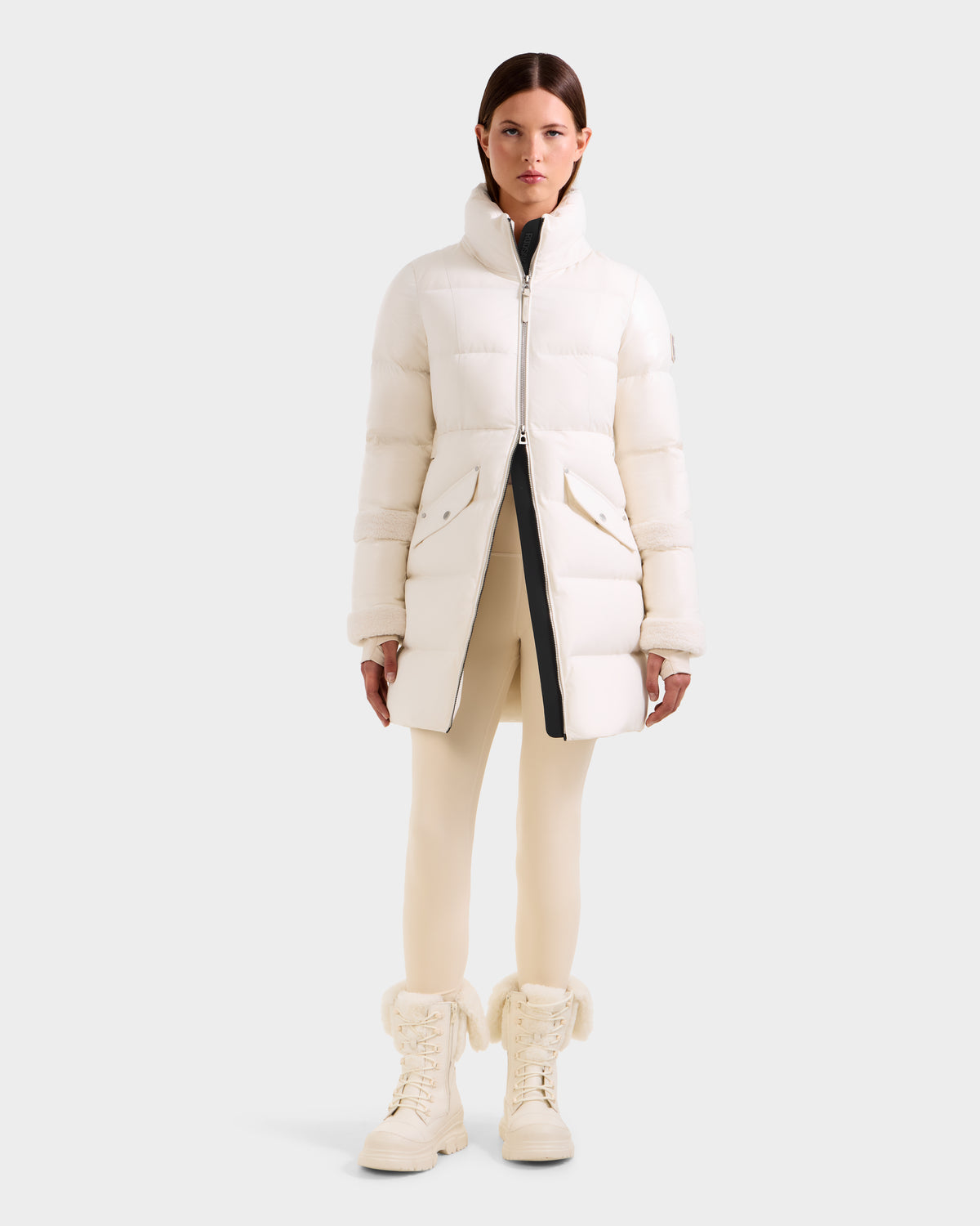 A woman faces forward wearing the Rudsak AUDREY L - 8124585 Vanilla knee-length down puffer jacket, cream leggings, and matching fur-trimmed lace-up boots against a plain light gray background.
