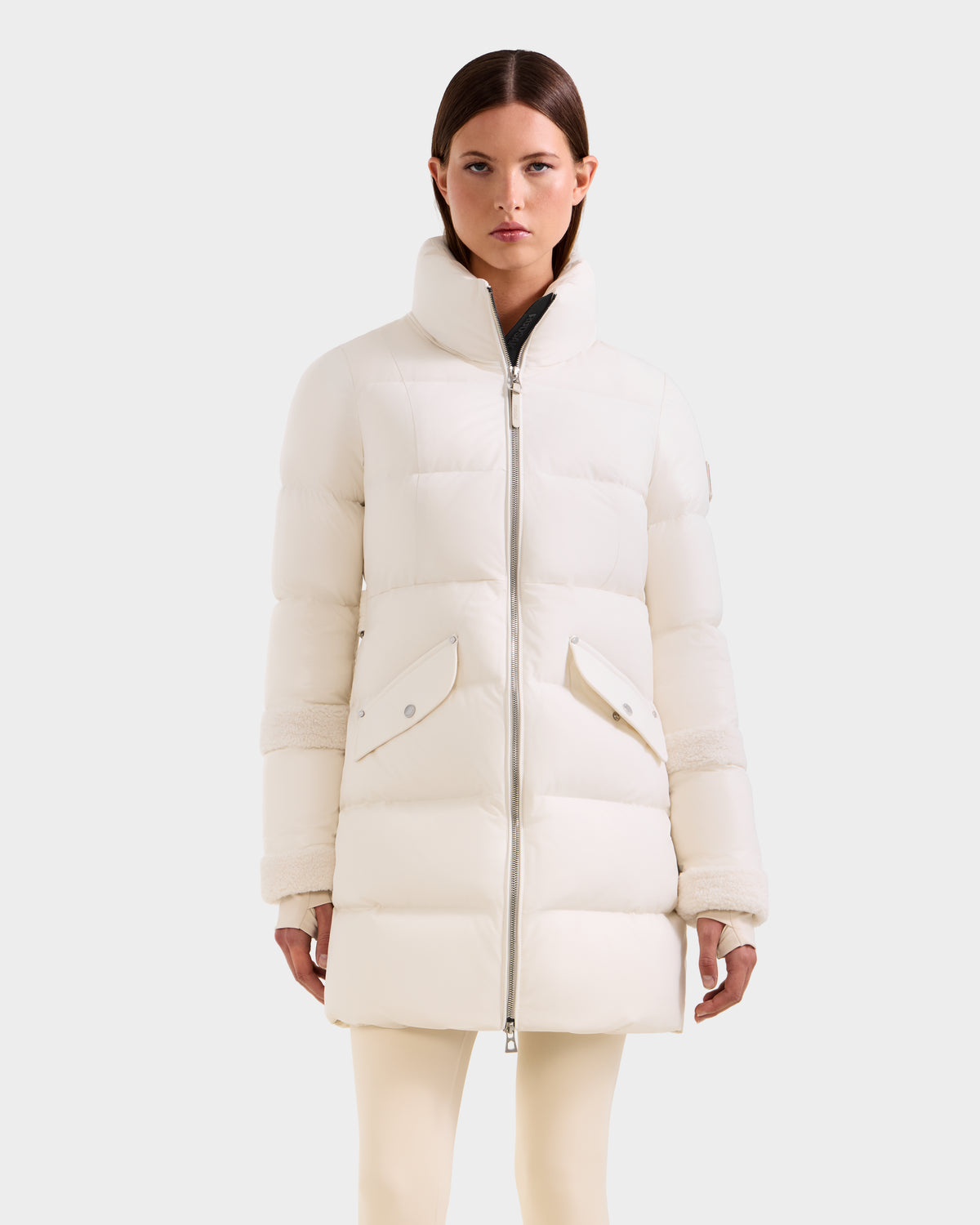 A woman with straight brown hair stands facing forward against a light background, wearing the Rudsak AUDREY L - 8124585 Vanilla down puffer jacket made from recycled Stormshell fabric with a high collar, front zipper, and large flap pockets.