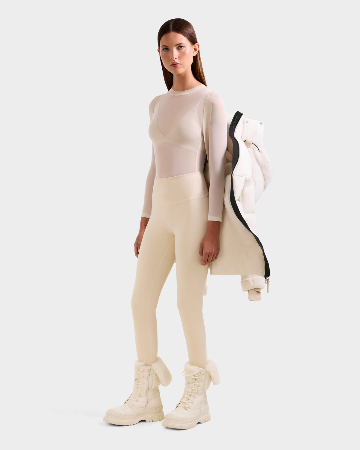 A woman poses in Rudsak’s AUDREY L - 8124585 Vanilla, a knee-length down puffer jacket crafted from recycled Stormshell fabric, draped over her shoulder against a plain background.