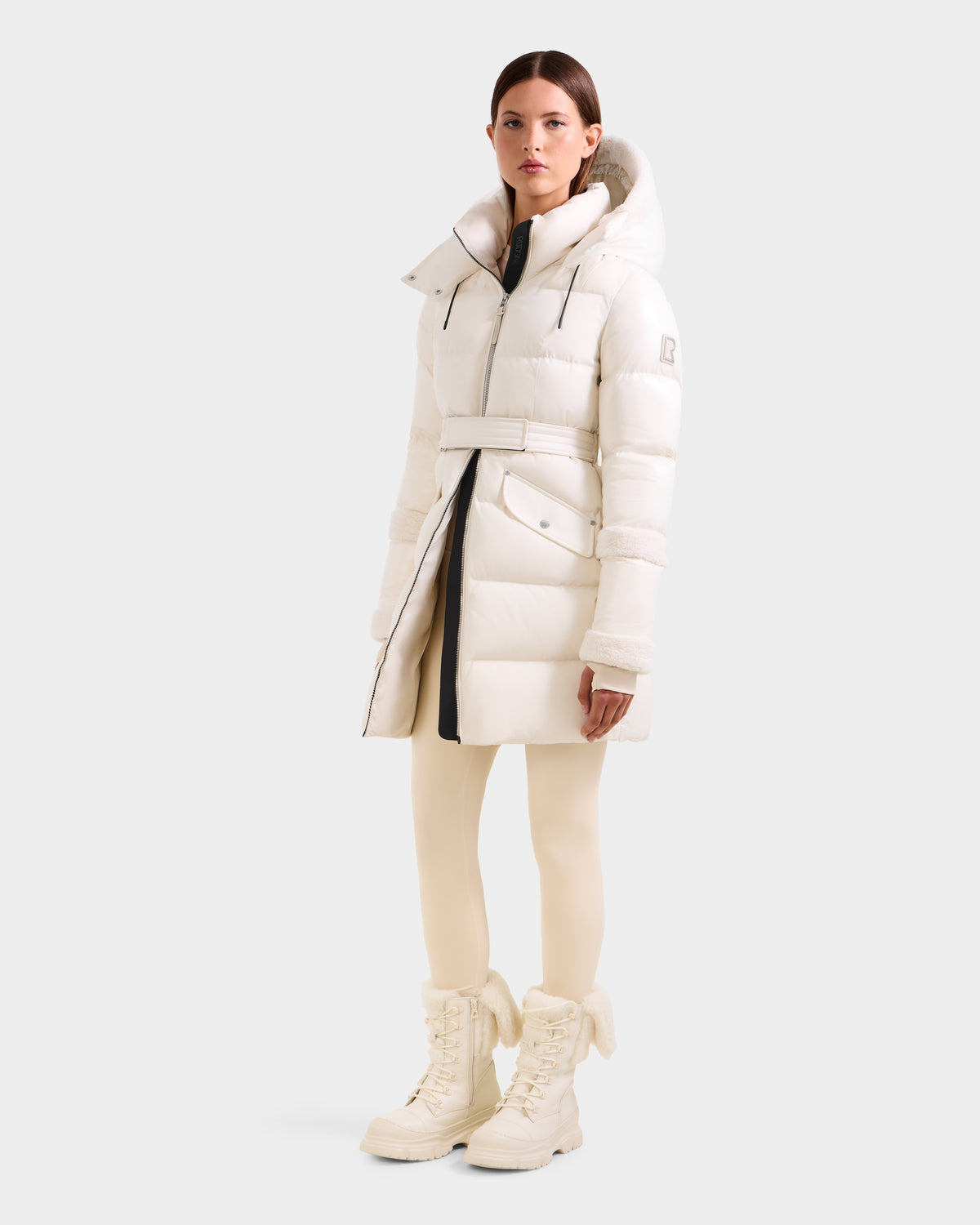 A woman poses against a light background wearing the Rudsak AUDREY L - 8124585 Vanilla belted knee-length down puffer with recycled Stormshell, hood, zip pockets, cream leggings, and matching lace-up boots.