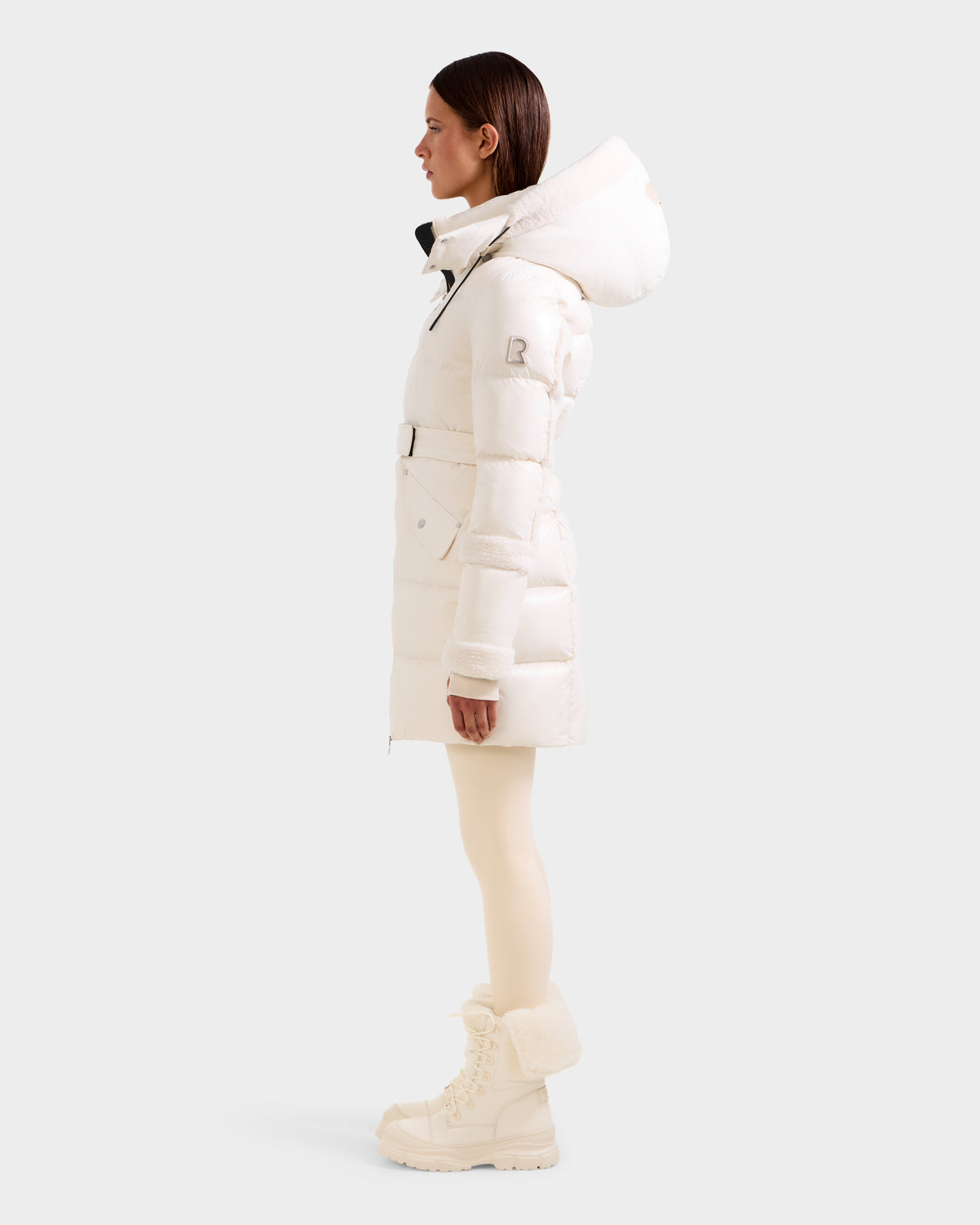 A woman stands in profile wearing the Rudsak AUDREY L - 8124585 Vanilla knee-length down puffer jacket with a hood, paired with cream tights and winter boots against a plain light background.