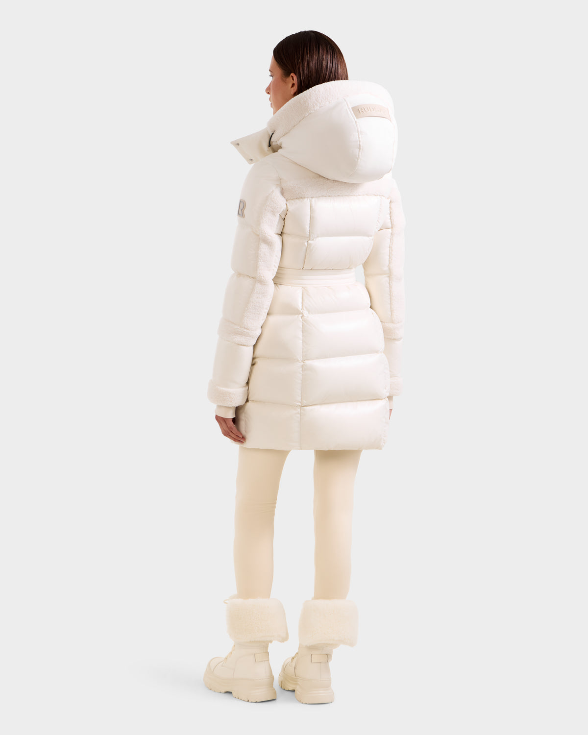 A woman faces away, wearing the Rudsak AUDREY L - 8124585 Vanilla off-white, knee-length down puffer jacket with a hood—crafted from recycled Stormshell fabric—against a light gray background.
