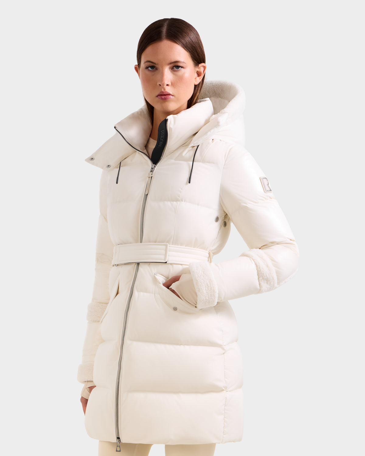 A woman with straight brown hair and a neutral expression models the Rudsak AUDREY L - 8124585 Vanilla: a long, white belted down puffer jacket with high collar and hood, crafted from recycled Stormshell, against a plain light background.