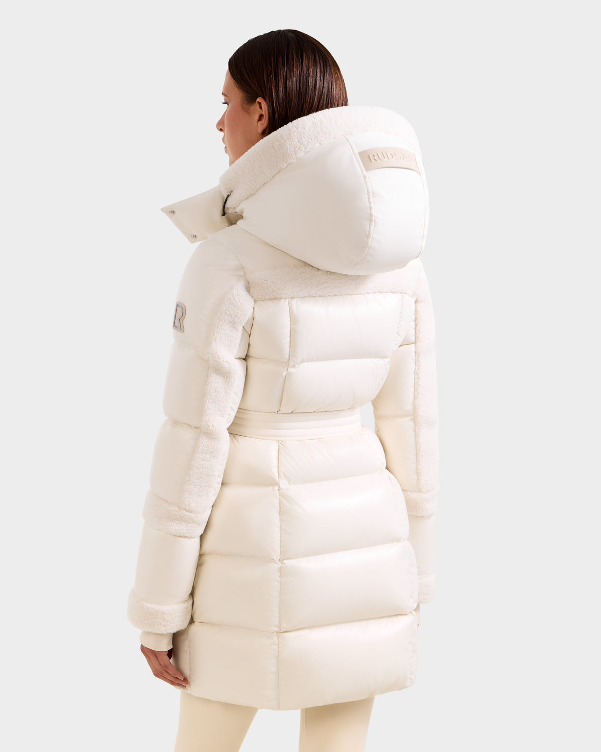 A woman with straight brown hair wears the Rudsak AUDREY L - 8124585 Vanilla cream down puffer jacket, featuring a large hood and textured side panels. Made with RDS certified down, she is shown from the back against a plain light background.