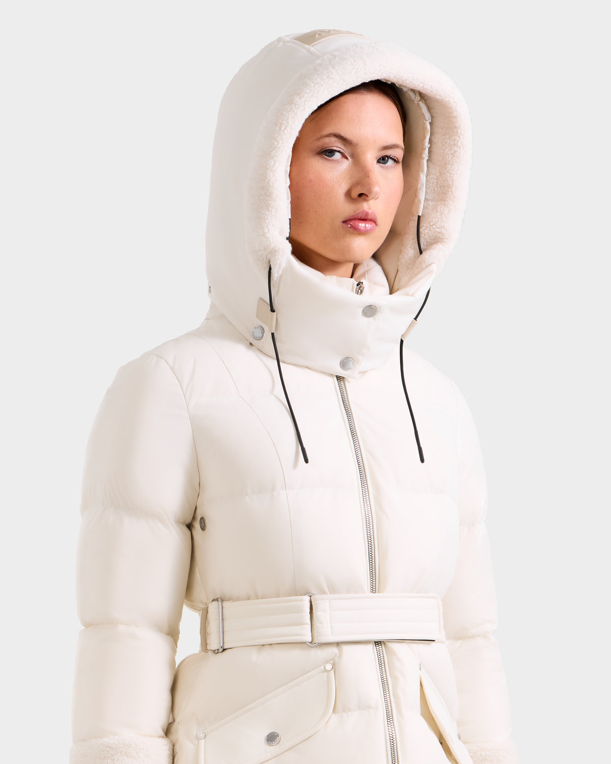 A woman models the Rudsak AUDREY L - 8124585 Vanilla: a knee-length puffer jacket with belted waist and faux fur-trimmed hood, made from recycled Stormshell fabric, standing against a plain light background.