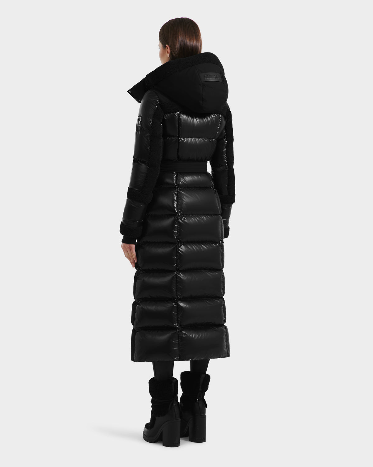 A woman faces away, wearing the Rudsak AUDREY XL - 8125601 Black/Black maxi-length down puffer jacket with a large hood and black heeled boots. Her straight brown hair is visible, set against a plain white background.