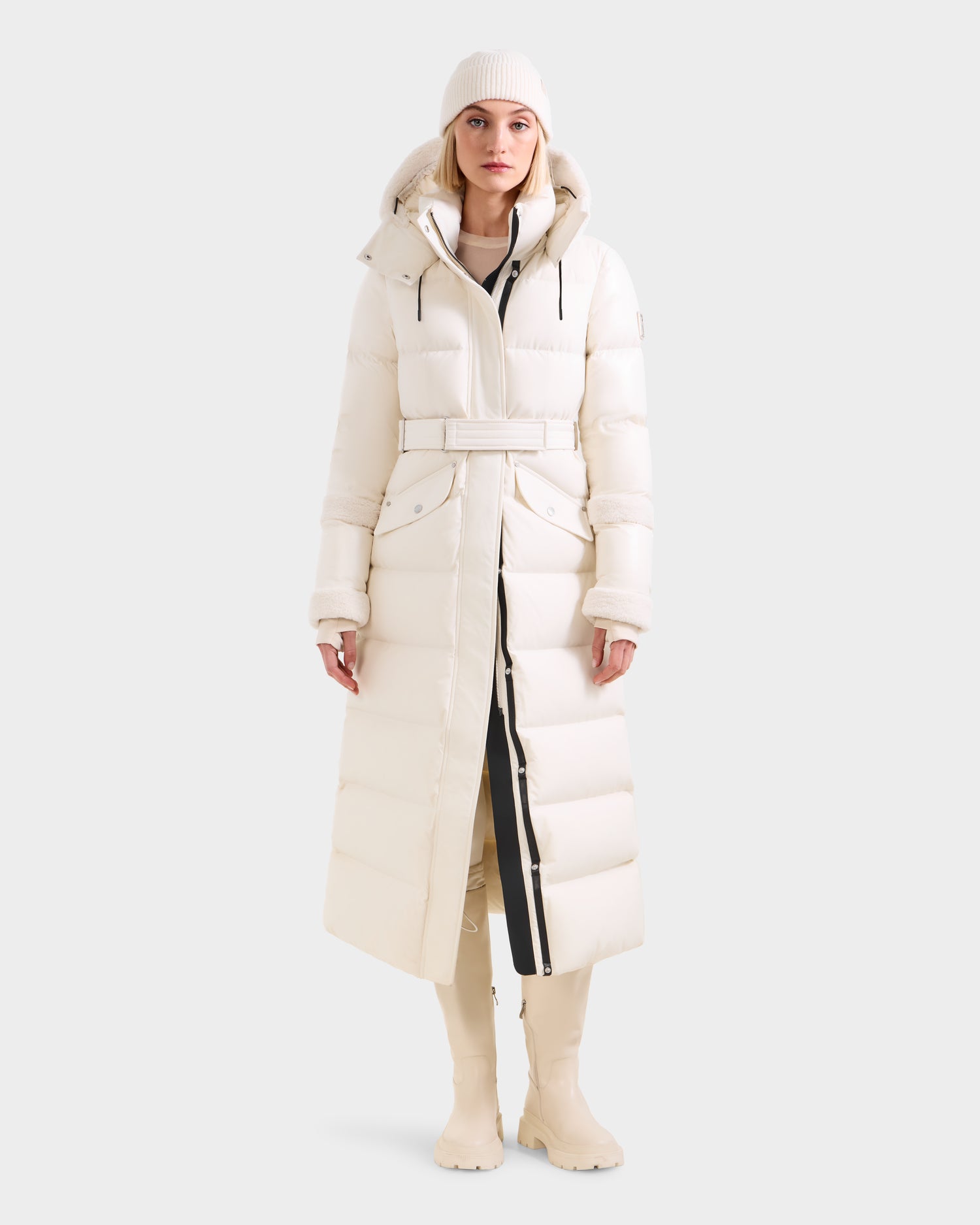 A woman models the Rudsak AUDREY XL - 8125601 Vanilla maxi-length down puffer jacket with belted waist and hood, styled with a white knit beanie and knee-high boots, standing against a plain light background.