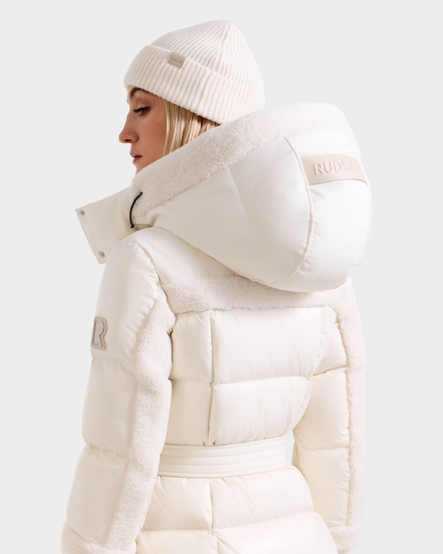 A person wears the Rudsak AUDREY XL - 8125601 Vanilla white maxi down puffer with textured panels, recycled Stormshell fabric, and a RUDSAK patch on the hood, paired with a white knit beanie. Seen from the back, slightly turned to the side.
