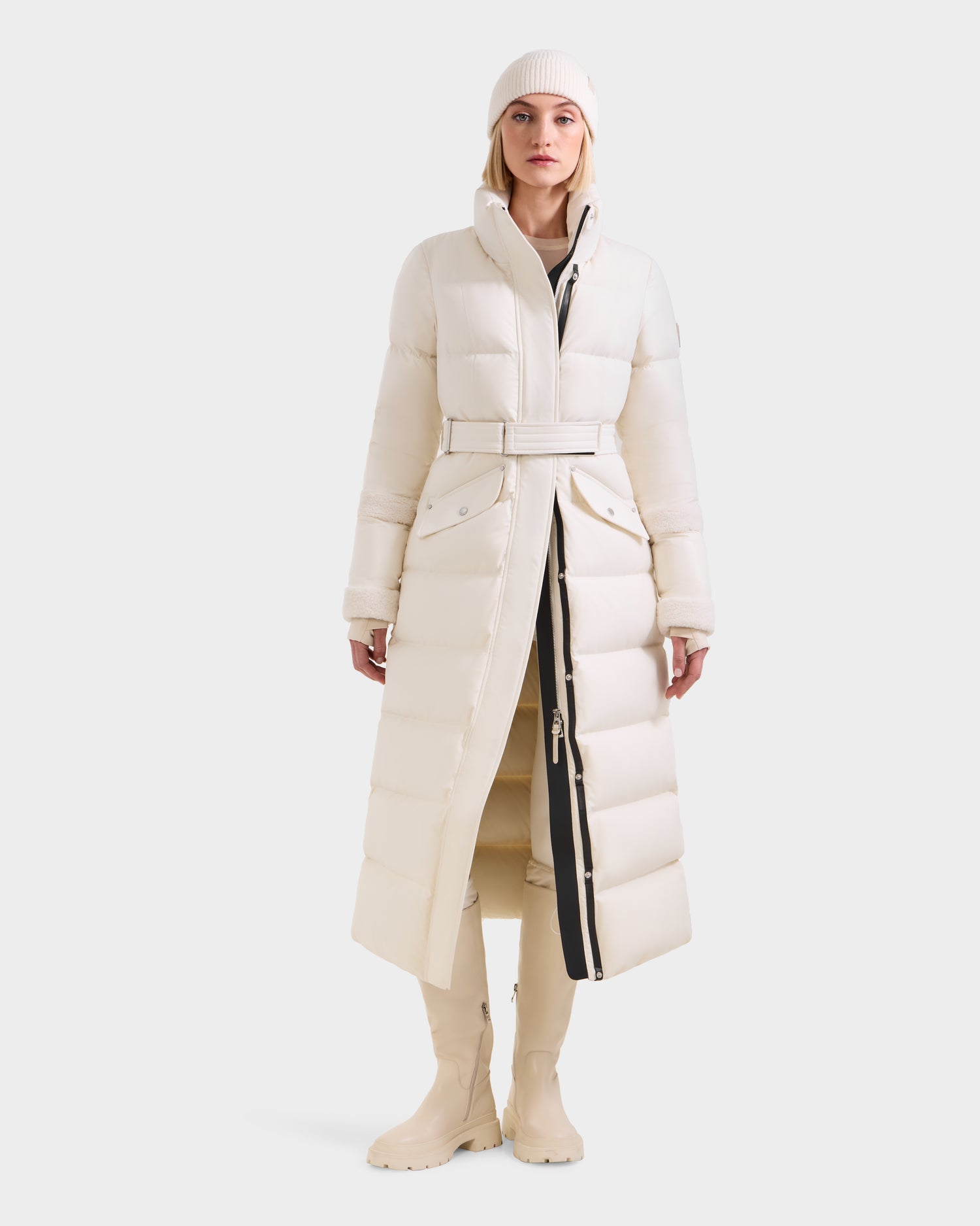 A woman wears the Rudsak AUDREY XL - 8125601 Vanilla maxi down puffer in white recycled Stormshell, belted at the waist, paired with a matching beanie and knee-high boots. The coat is buttoned and zipped as she stands against a light background.