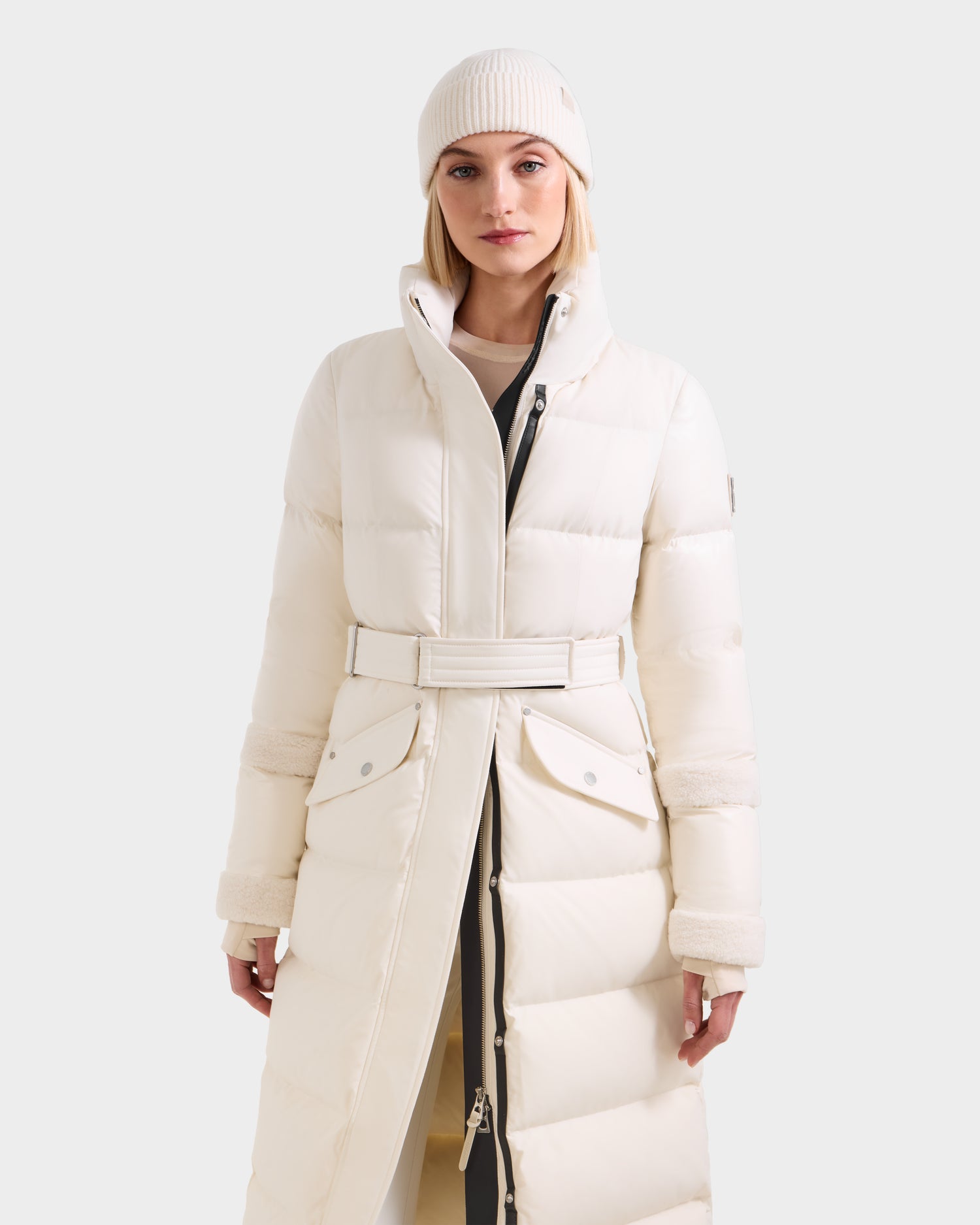 A woman in the Rudsak AUDREY XL - 8125601 Vanilla maxi-length down puffer jacket and a white knit beanie stands facing forward against a plain, light background.