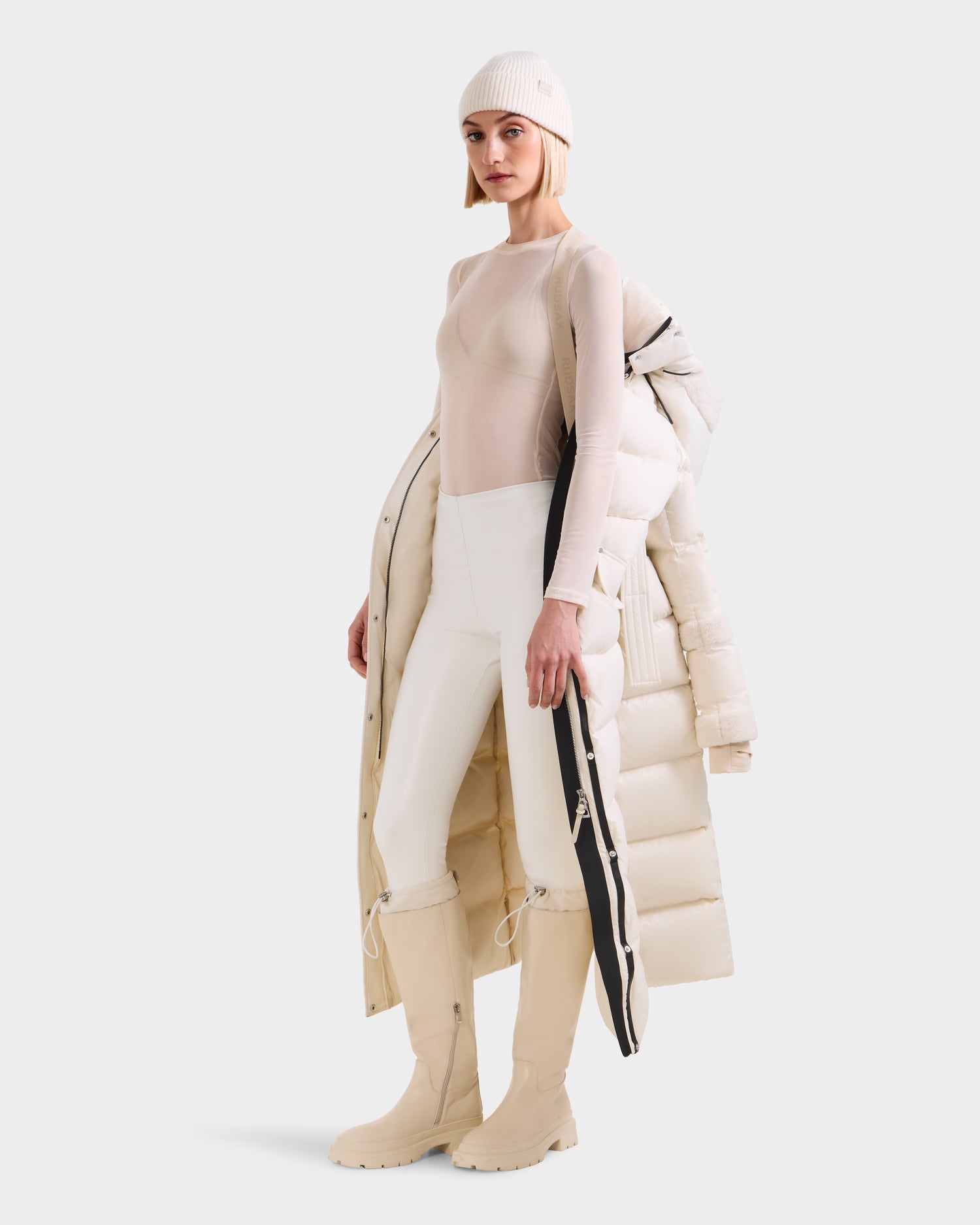 A woman stands against a plain white background wearing the Rudsak AUDREY XL - 8125601 Vanilla maxi-length down puffer jacket with faux shearling trim, styled over one shoulder, paired with a beige beanie, sheer top, white pants, and knee-high beige boots.