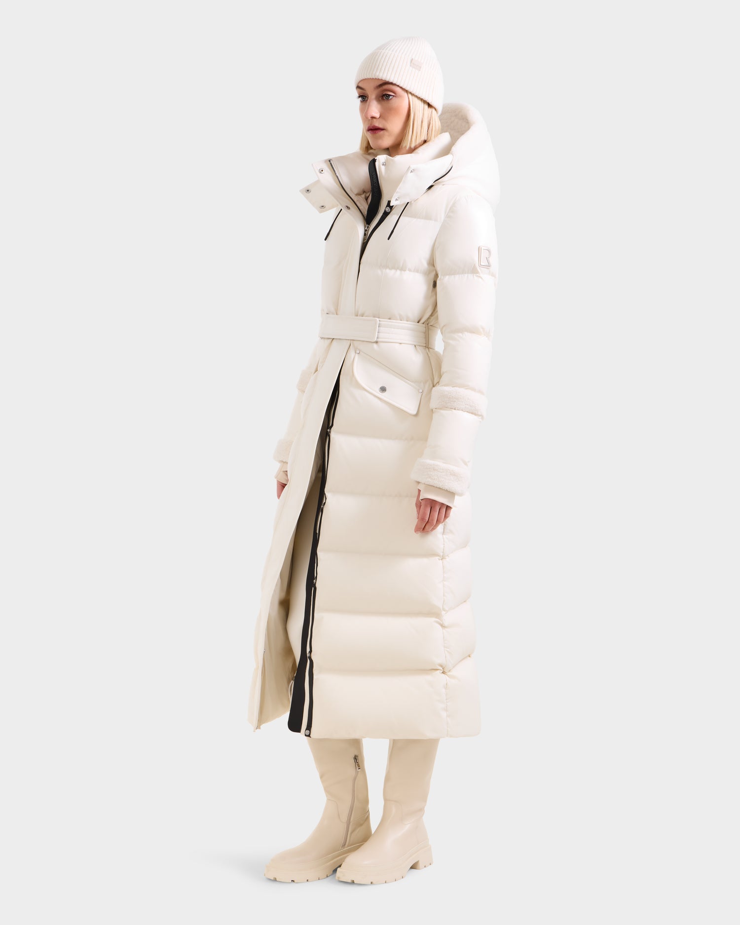 A woman wears the Rudsak AUDREY XL - 8125601 Vanilla maxi-length down puffer jacket with faux shearling trim, white boots, and a knit beanie, standing against a plain light background, facing slightly to the side.