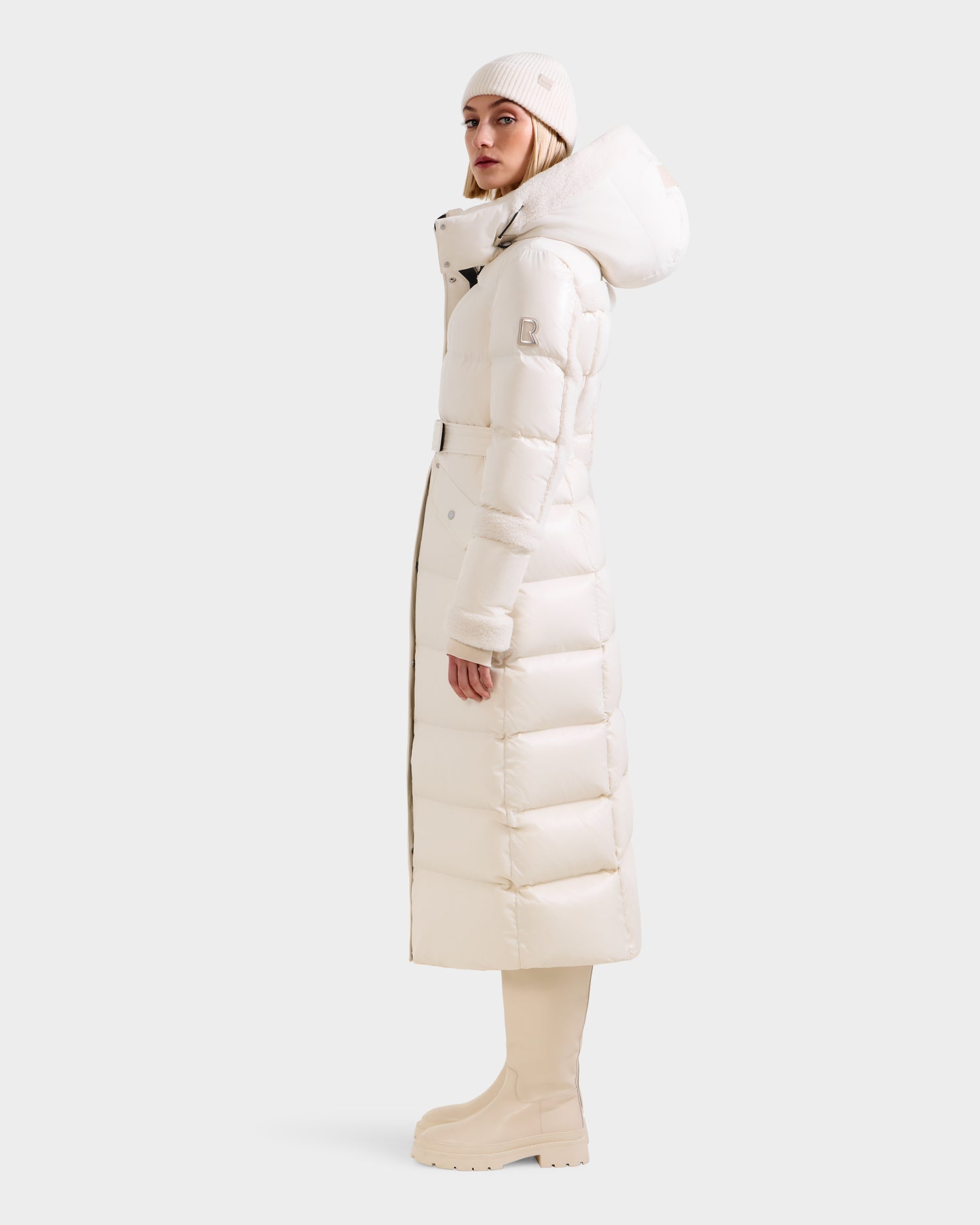 A woman stands sideways in a Rudsak AUDREY XL - 8125601 Vanilla maxi-length down puffer jacket with a hood, paired with a white knit beanie, tall boots, and light pants against a plain light background.