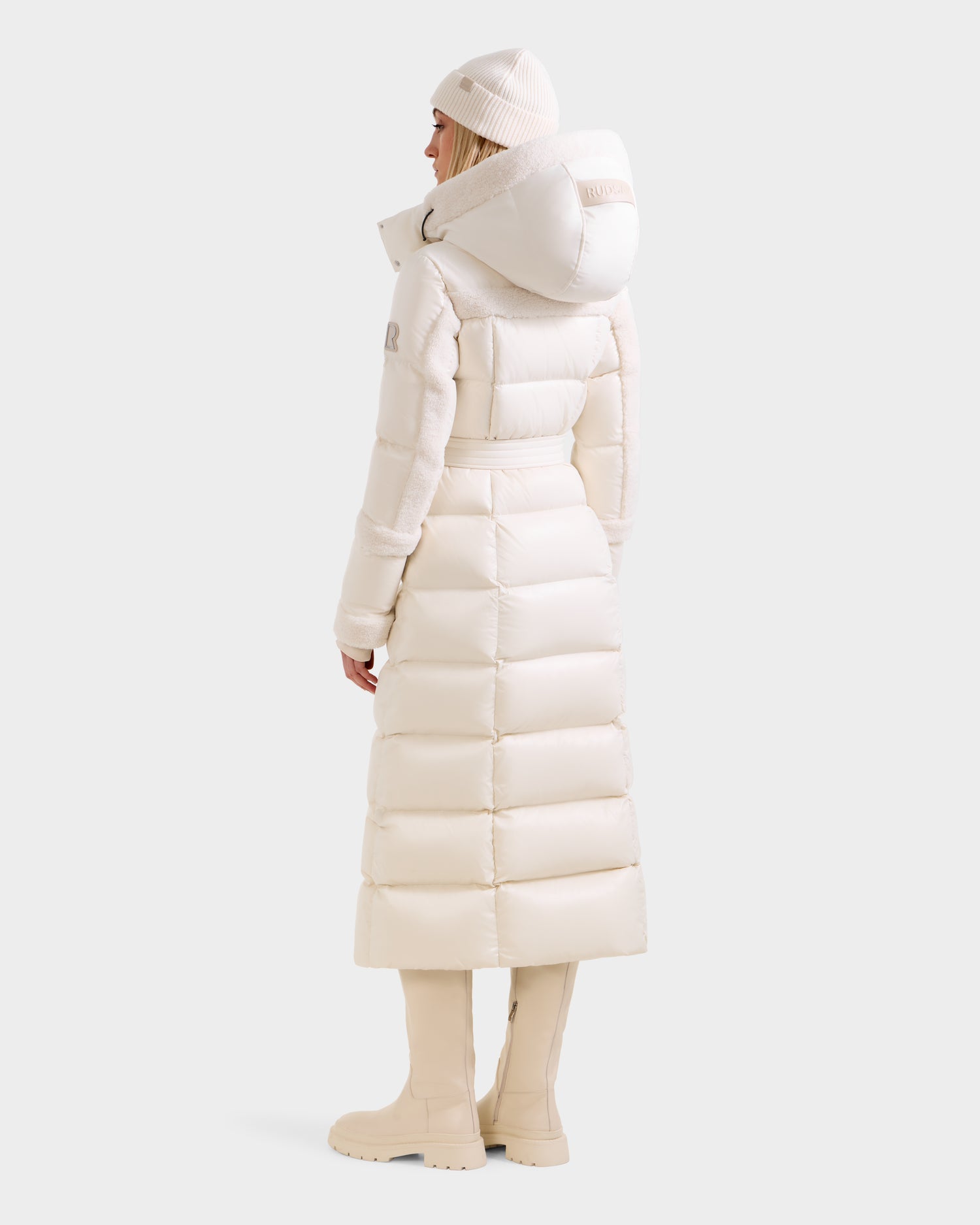 A woman models the Rudsak AUDREY XL - 8125601 Vanilla maxi-length down puffer, paired with a matching knit hat and tall boots, shown from the back against a plain light background.