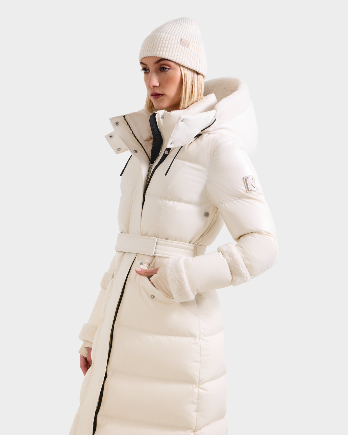 Wearing the Rudsak AUDREY XL - 8125601 Vanilla, a maxi-length down puffer with recycled Stormshell fabric, high collar, hood, and belt, she pairs it with a cream knit beanie and stands against a plain light background.
