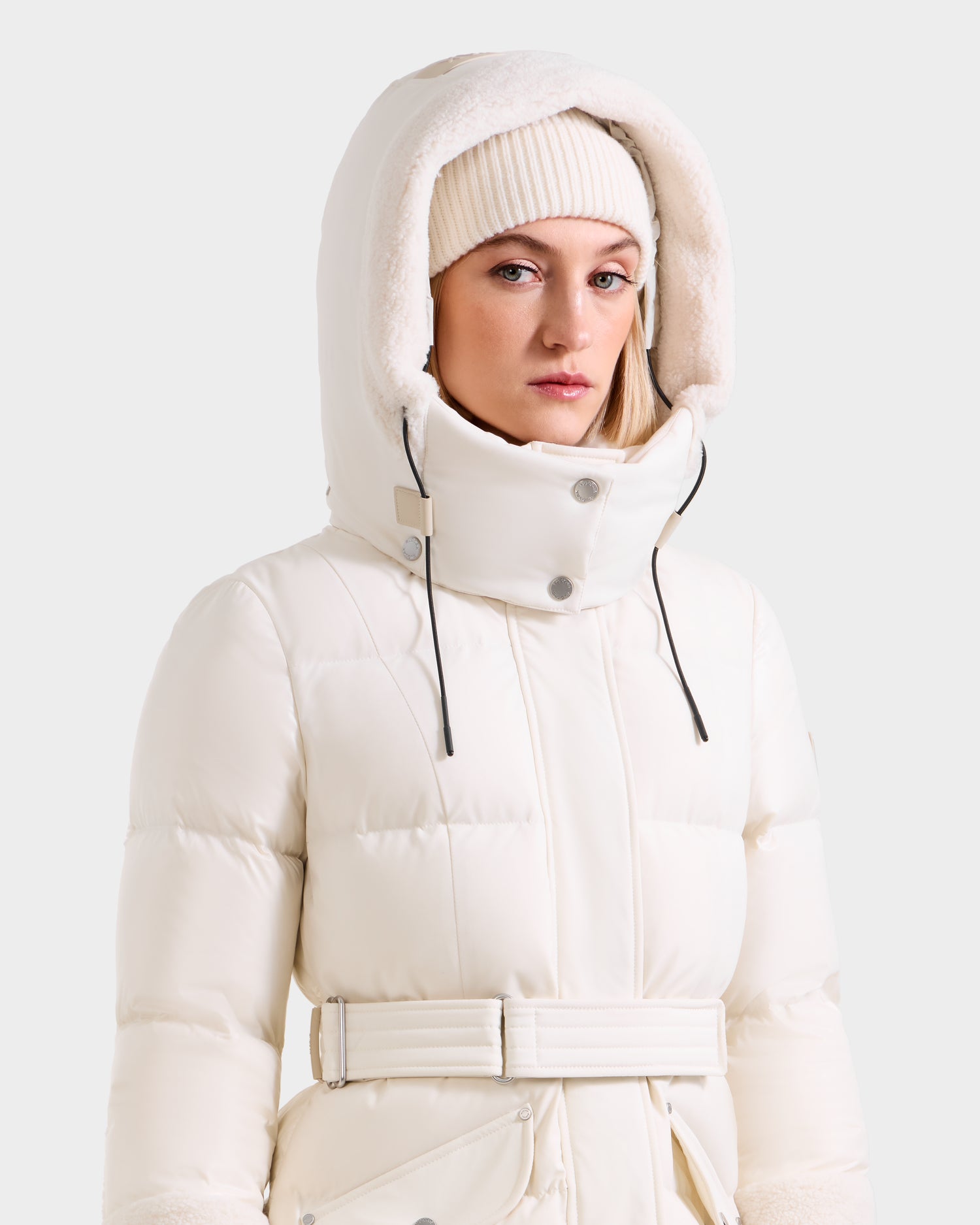 A woman wears the Rudsak AUDREY XL - 8125601 Vanilla maxi down puffer jacket with a belted waist and faux shearling trim over a white knit beanie, standing against a plain light background.