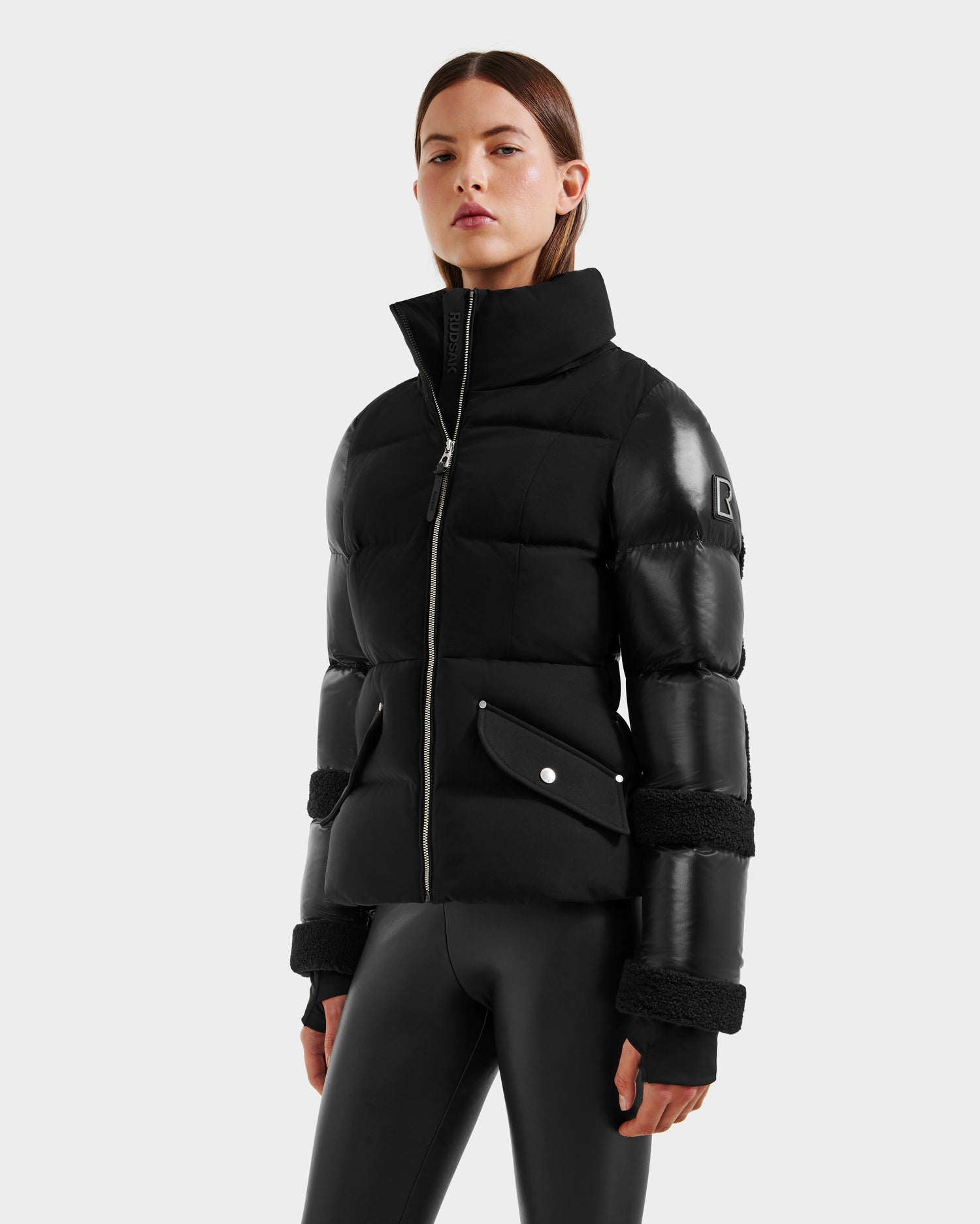 A person wears the AUDREY - 8124582 Black/Black down puffer jacket by Rudsak, featuring shiny leather accents and zippered front, crafted from recycled Stormshell fabric, paired with black leggings against a plain light background.