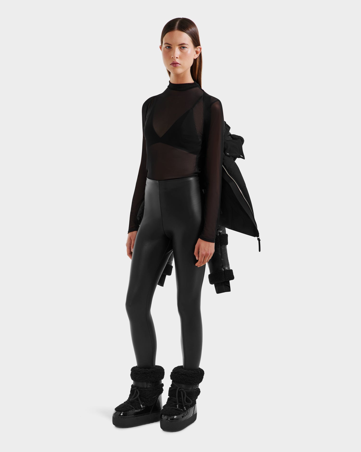A woman stands against a plain background wearing the Rudsak AUDREY - 8124582 Black/Black recycled Stormshell jacket draped over her shoulders, paired with a sheer black long-sleeve top, black bra, high-waisted leggings, and chunky boots.