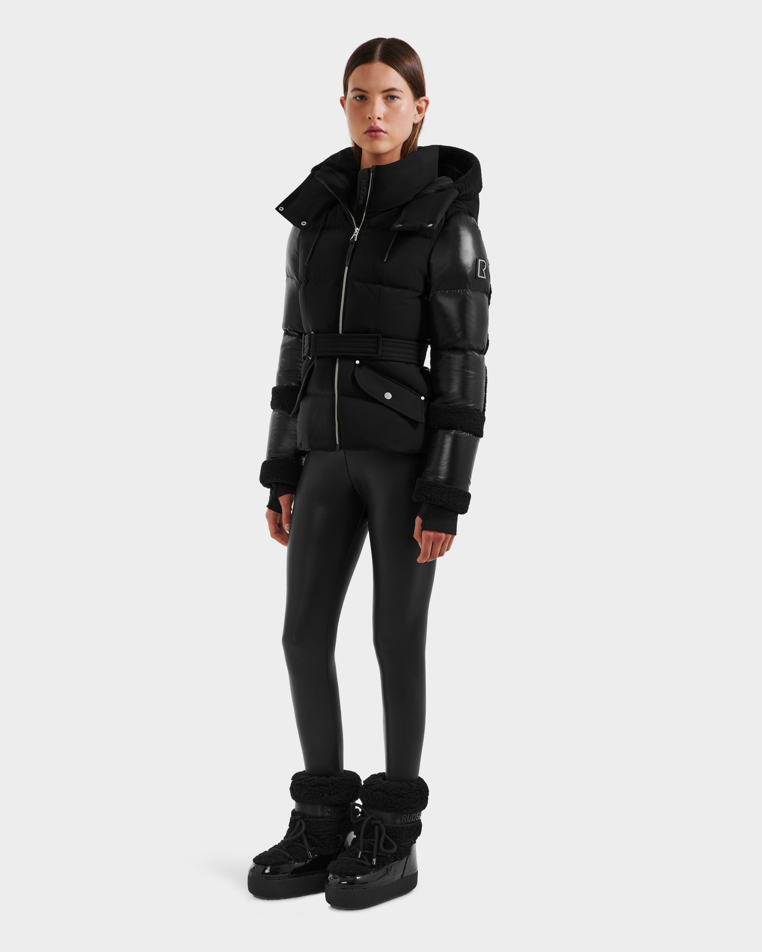 A woman stands against a plain light gray background wearing the Rudsak AUDREY - 8124582 Black/Black down puffer jacket with high collar and belt, black shiny leggings, and black fur-trimmed boots.