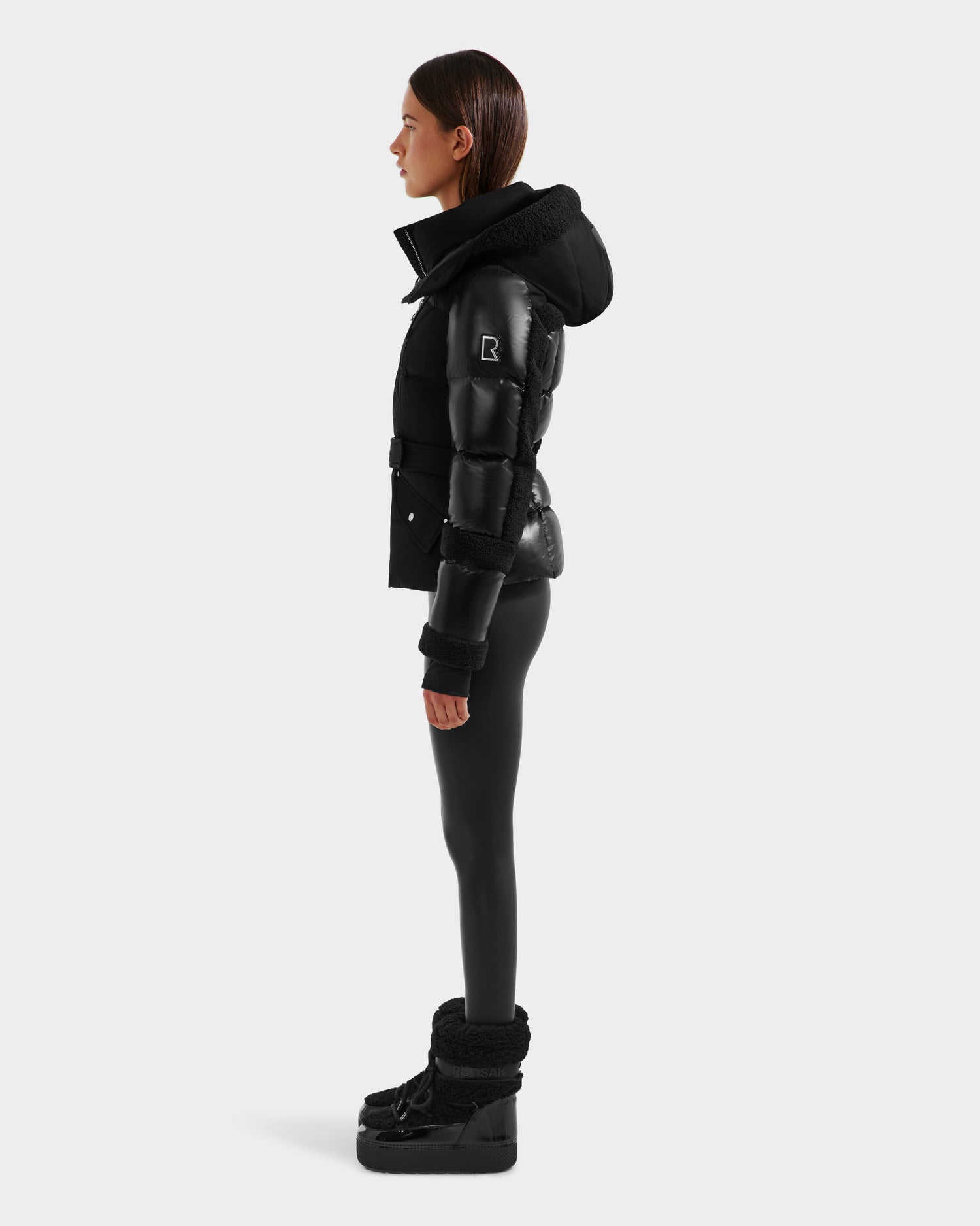 A woman stands in profile against a plain background, wearing the Rudsak AUDREY - 8124582 Black/Black down puffer jacket, black leggings, and black winter boots. Her arms rest by her sides and her straight dark hair is shoulder-length.