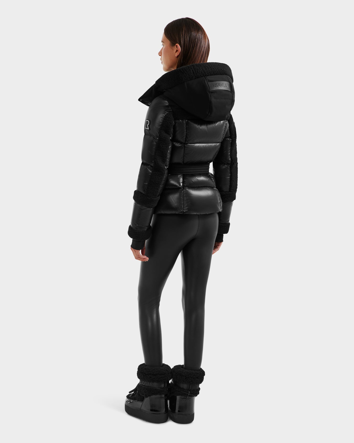 A woman with straight brown hair faces away, wearing the Rudsak AUDREY - 8124582 Black/Black shiny down puffer jacket made with recycled Stormshell fabric, black leggings, and black boots against a plain light background.
