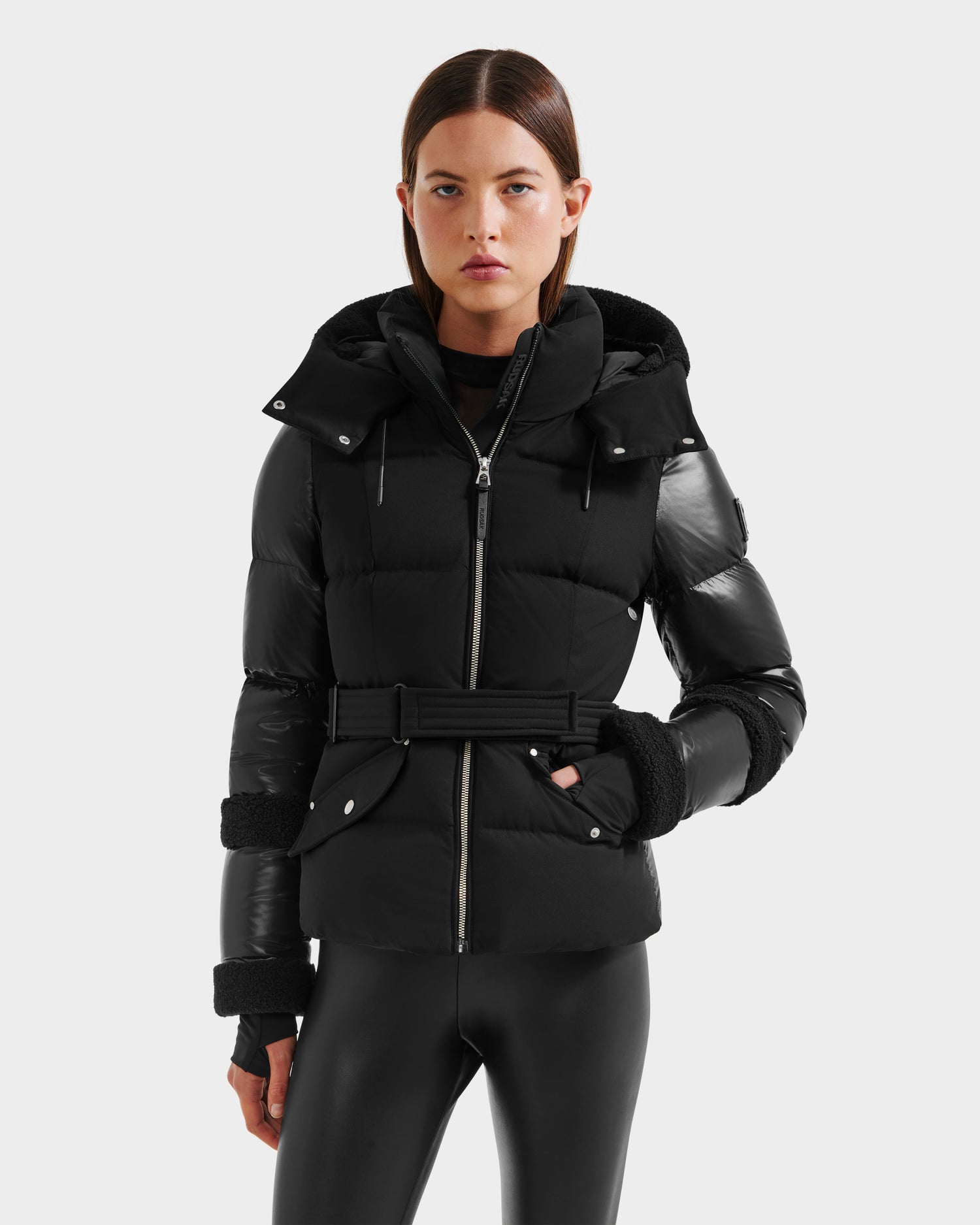 A woman with straight brown hair and a neutral expression wears the Rudsak AUDREY - 8124582 Black/Black fitted puffer jacket with RDS certified down, belted waist, black gloves, and shiny black leggings against a plain white background.