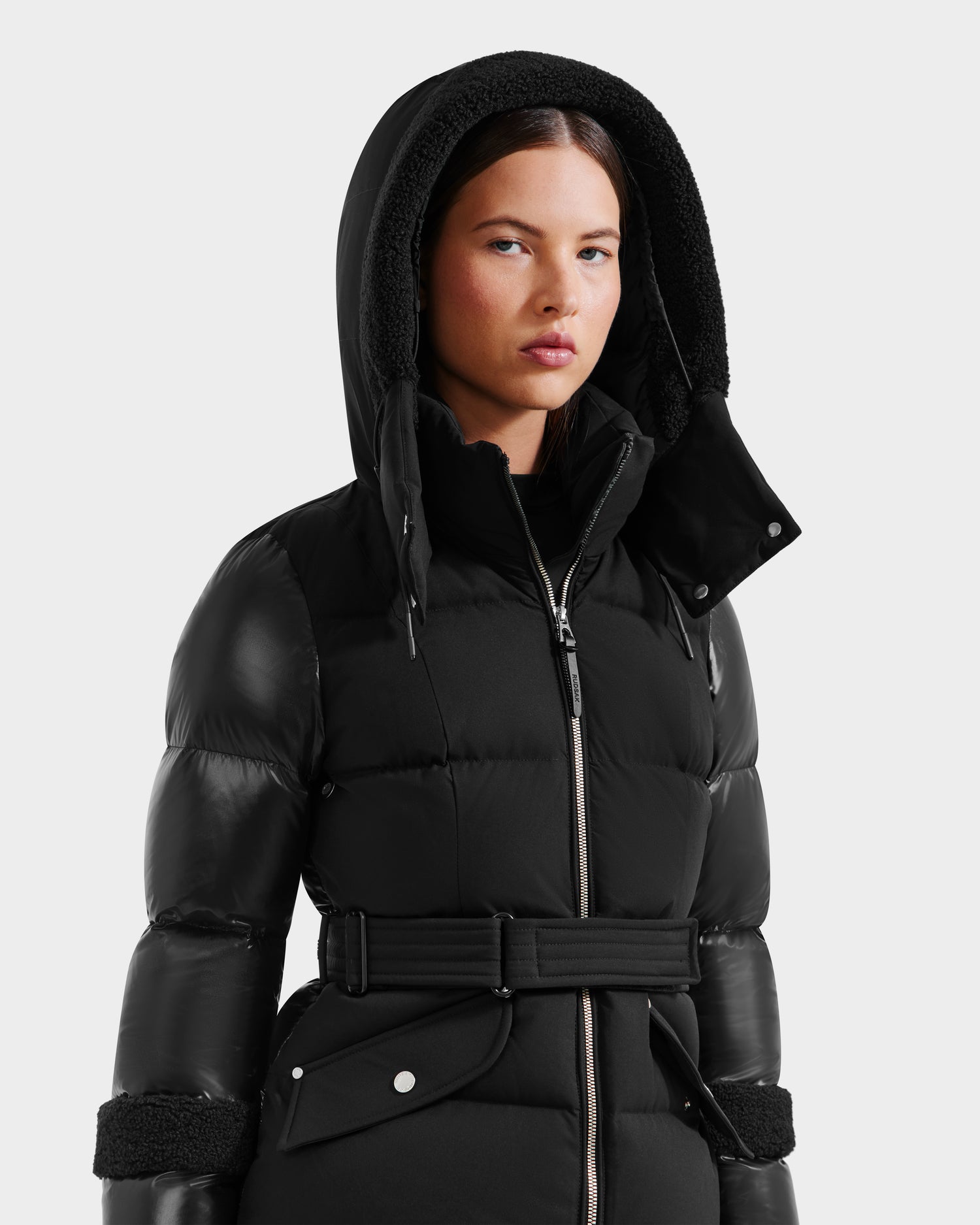 A woman wears the Rudsak AUDREY - 8124582 Black/Black down puffer jacket with RDS certified down, a belted waist, and faux fur-trimmed hood, looking forward with a neutral expression against a plain light background.