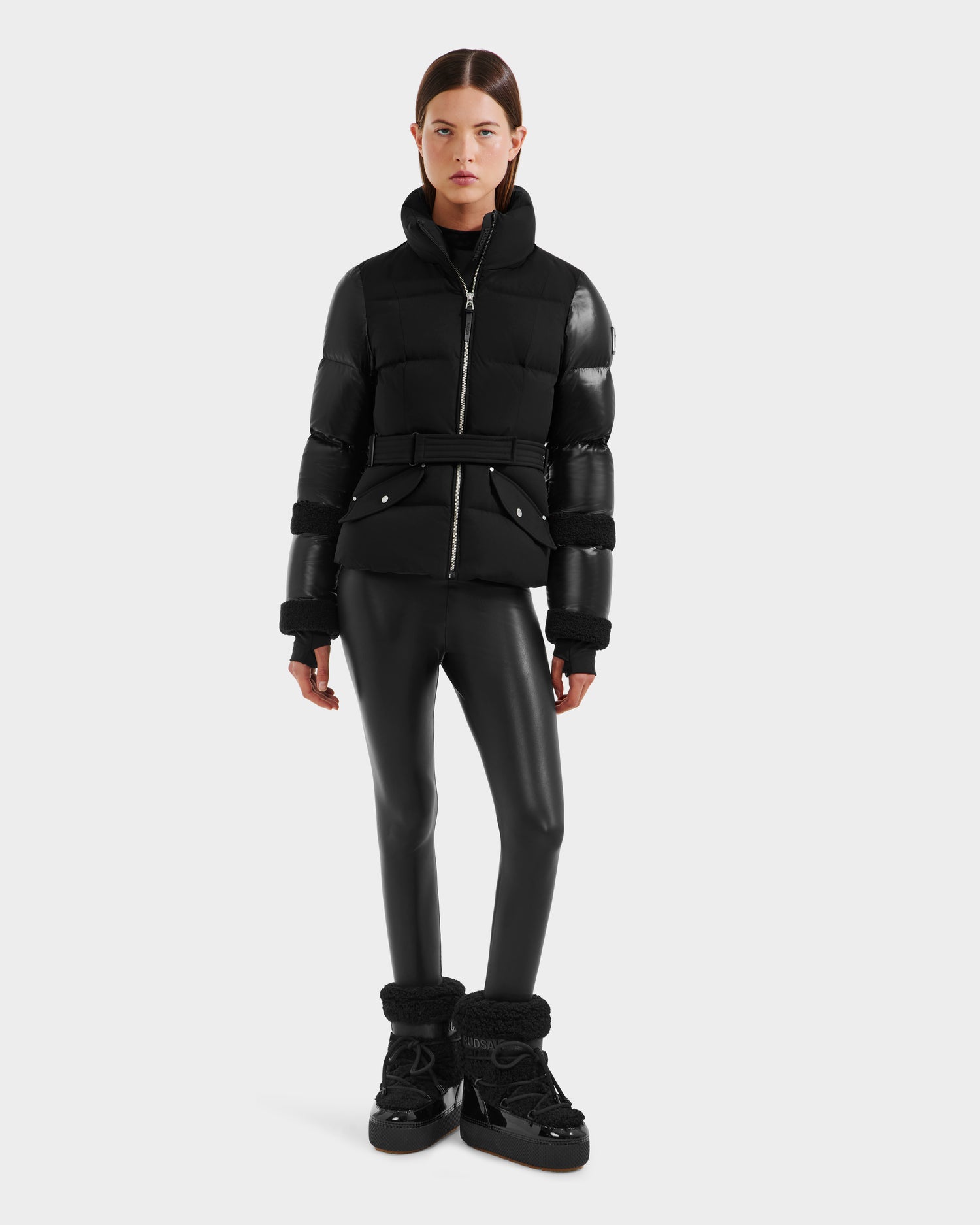 A woman faces forward in the Rudsak AUDREY - 8124582 Black/Black down puffer jacket made from recycled Stormshell, paired with black faux leather leggings and winter boots. She has straight dark hair and a neutral expression against a light background.
