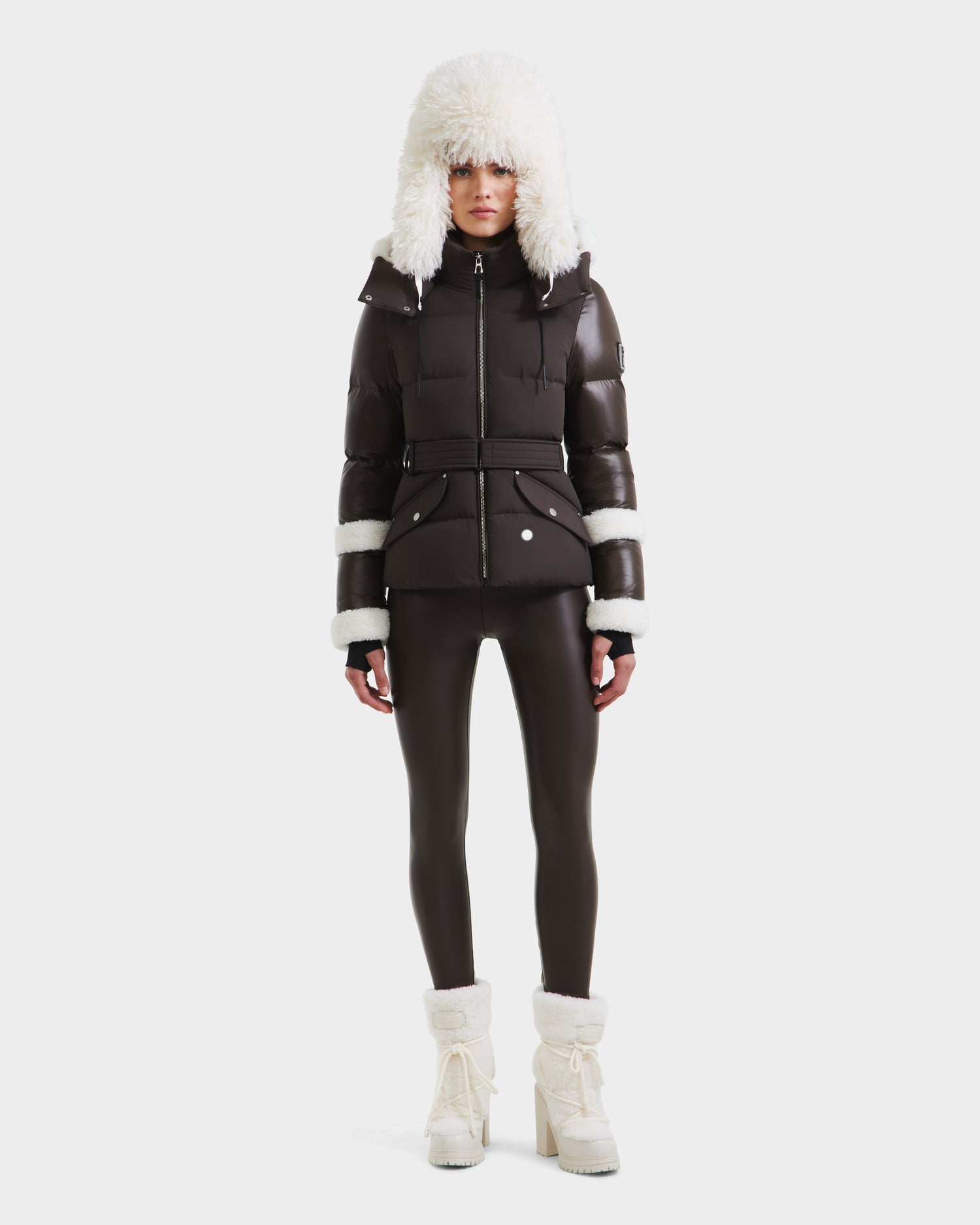 A woman wears the Rudsak AUDREY - 8124582 Espresso dark brown down puffer jacket with a belted waist, faux fur-trimmed hood and cuffs, matching leggings, and white heeled winter boots against a plain white background.