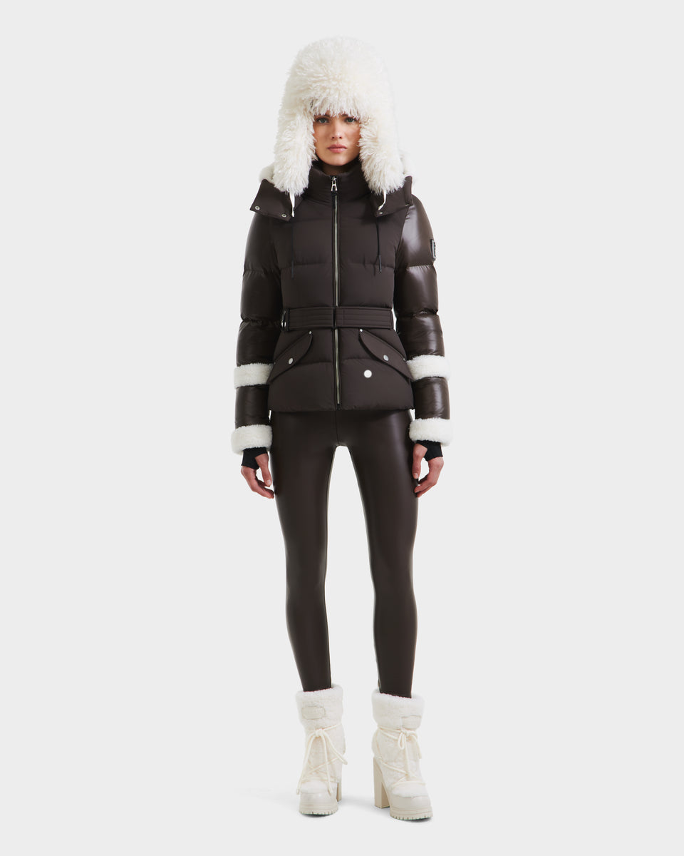 A woman wears the Rudsak AUDREY - 8124582 Espresso dark brown down puffer jacket with a belted waist, faux fur-trimmed hood and cuffs, matching leggings, and white heeled winter boots against a plain white background.